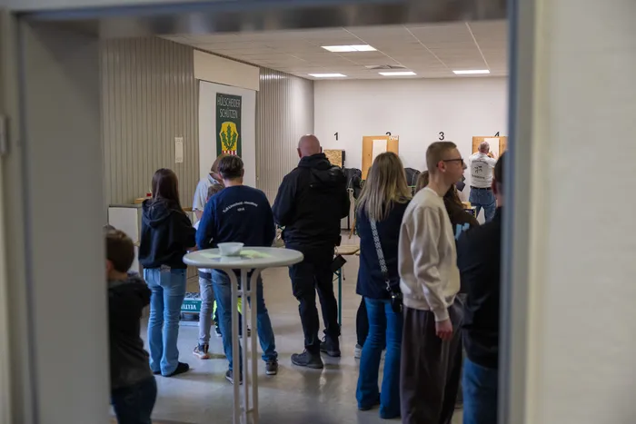 Menschen stehen in einer Schießhalle Schlange vor drei Schießständen, während andere zusehen. Im Hintergrund ein Banner mit der Aufschrift „Hülscheider Schützen“.