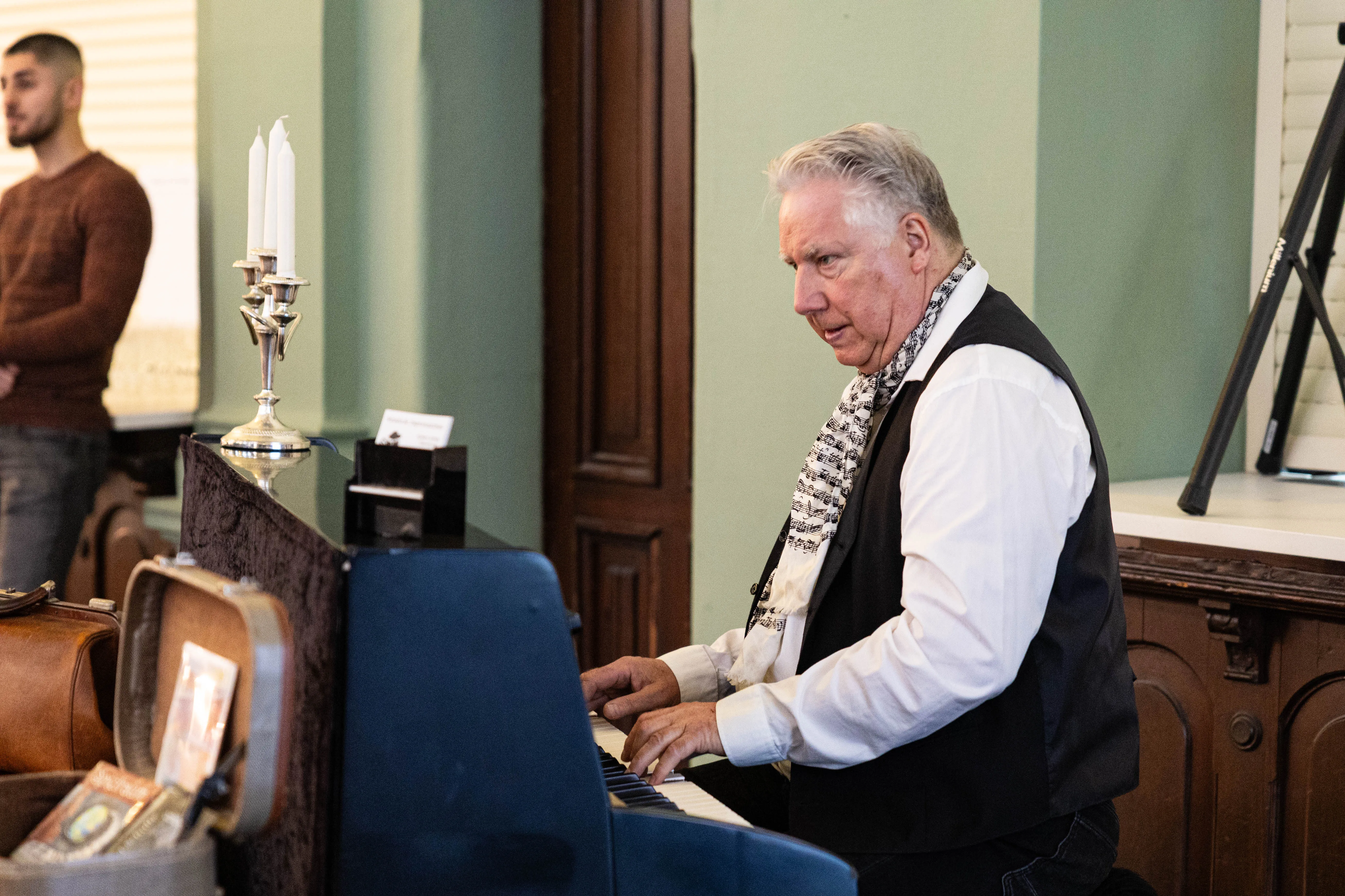 Ein älterer Mann spielt auf einem Klavier in einem Raum mit grünen Wänden. Im Hintergrund ist ein junger Mann zu sehen.