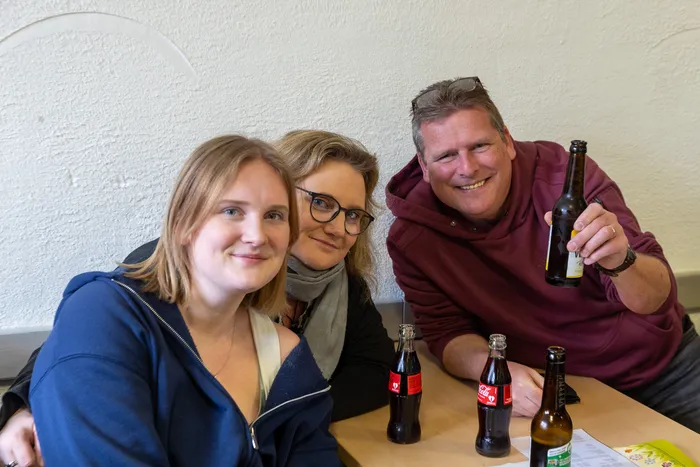 Drei lächelnde Menschen sitzen an einem Tisch mit Getränken, darunter Bierflaschen und Cola. Im Hintergrund ist eine helle Wand zu sehen.