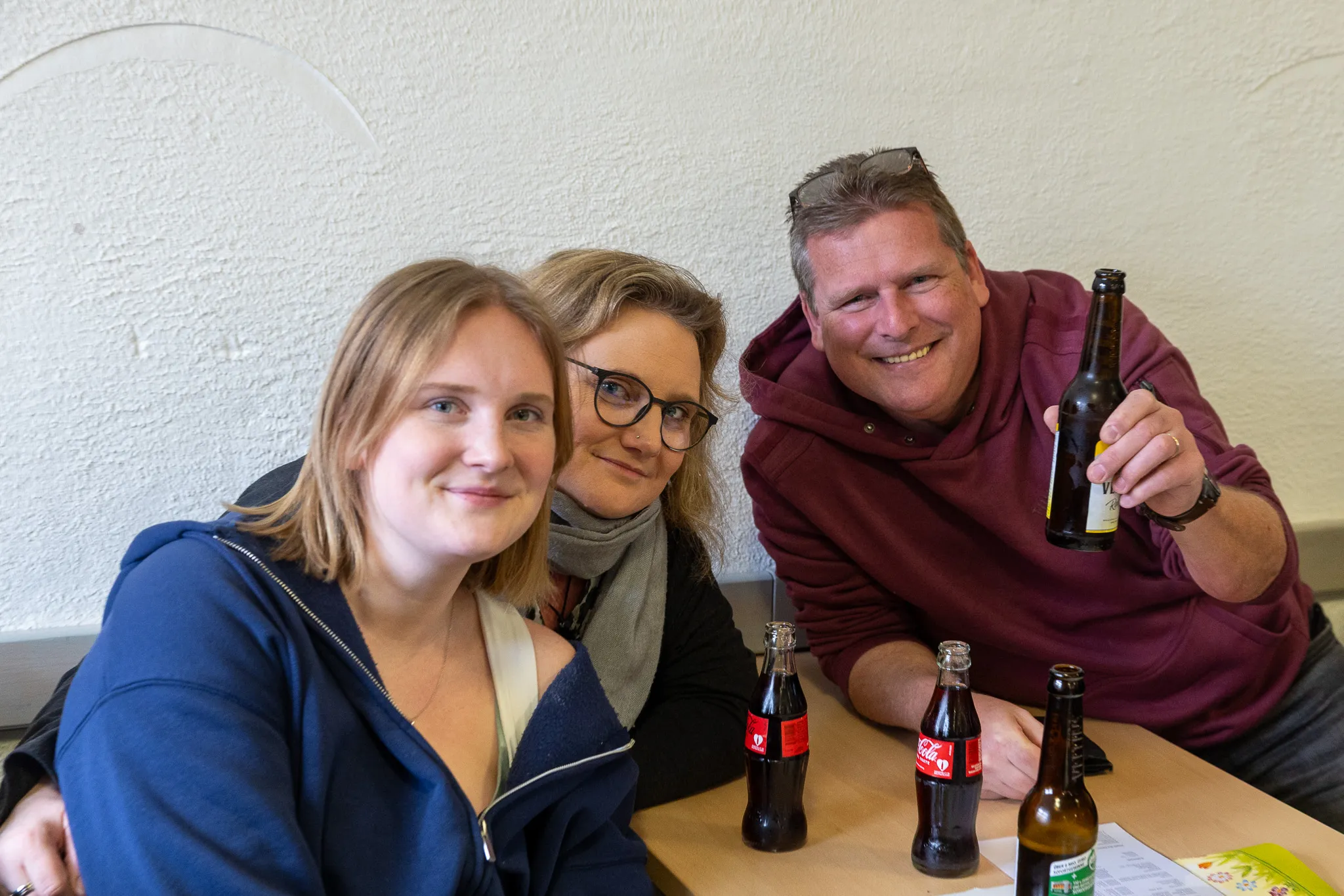 Drei lächelnde Menschen sitzen an einem Tisch mit Getränken, darunter Bierflaschen und Cola. Im Hintergrund ist eine helle Wand zu sehen.