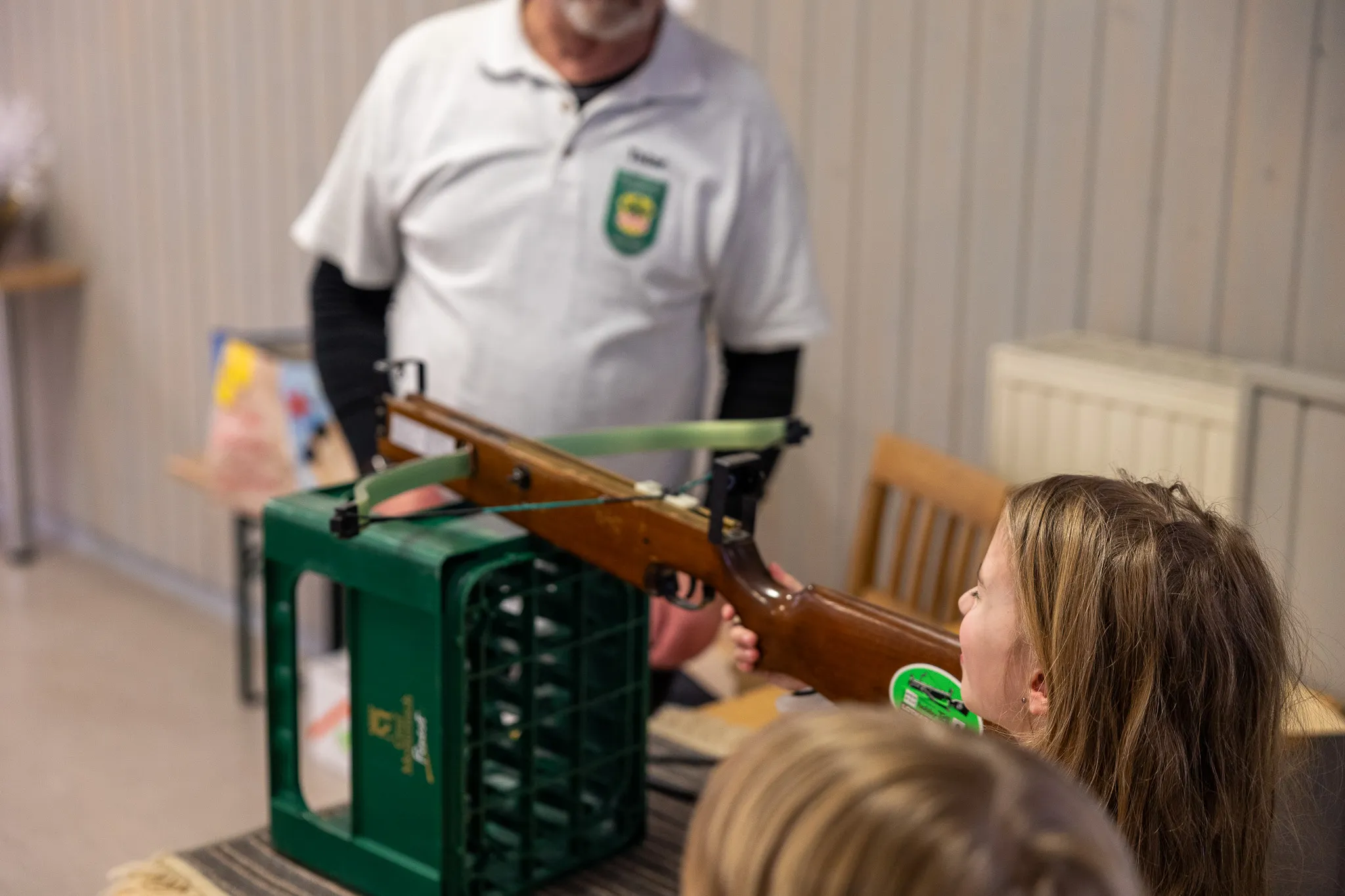 Ein Mädchen zielt mit einem Gewehr auf eine Zielscheibe, während ein Mann im Hintergrund beaufsichtigt. Das Gewehr ruht auf einer Kiste. Im Hintergrund ist ein Raum mit Heizkörpern zu sehen.