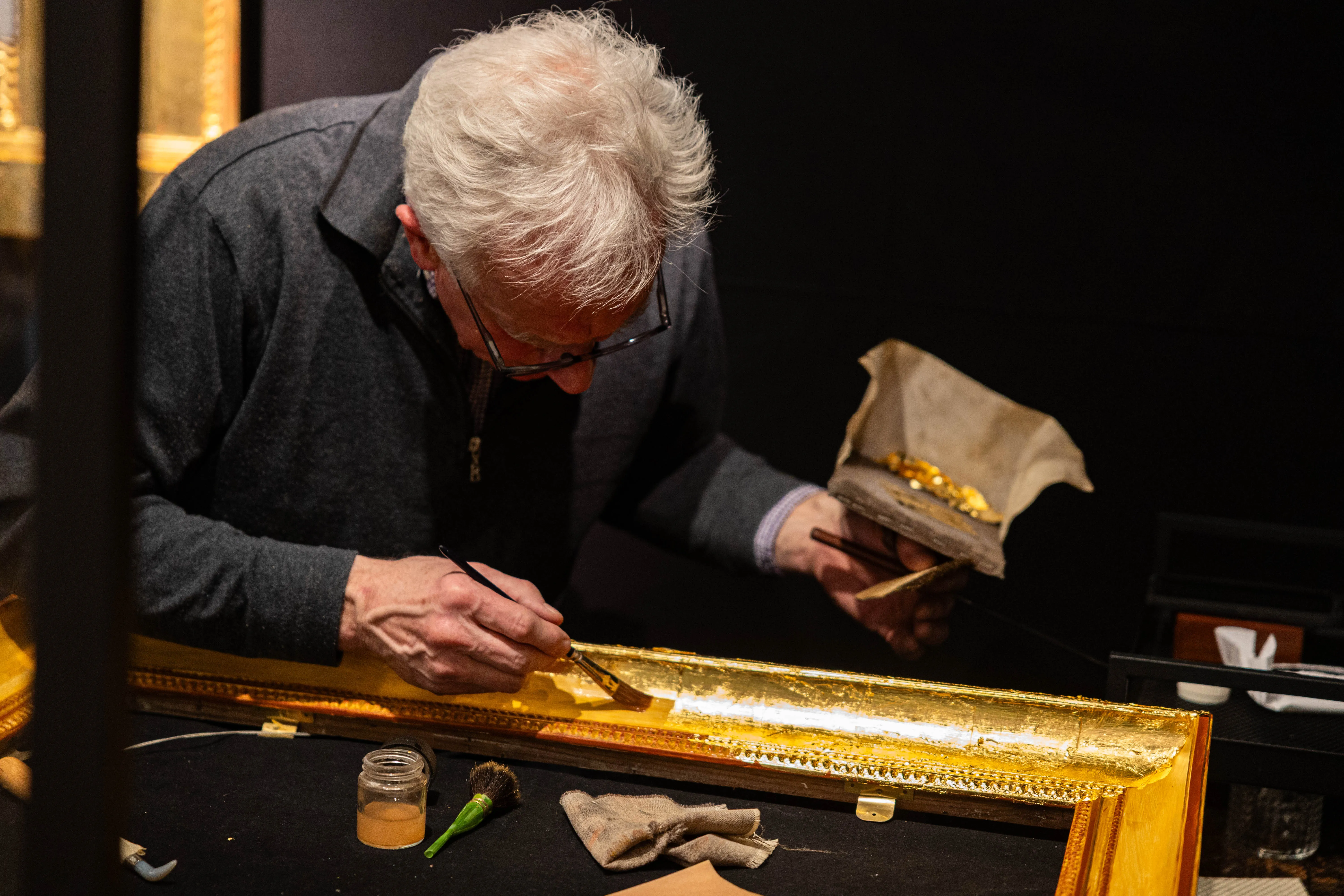 Ein Handwerker vergoldet einen kunstvollen Bilderrahmen mit einem feinen Pinsel. Neben ihm liegen Blattgold und Werkzeuge auf einer Werkbank.
