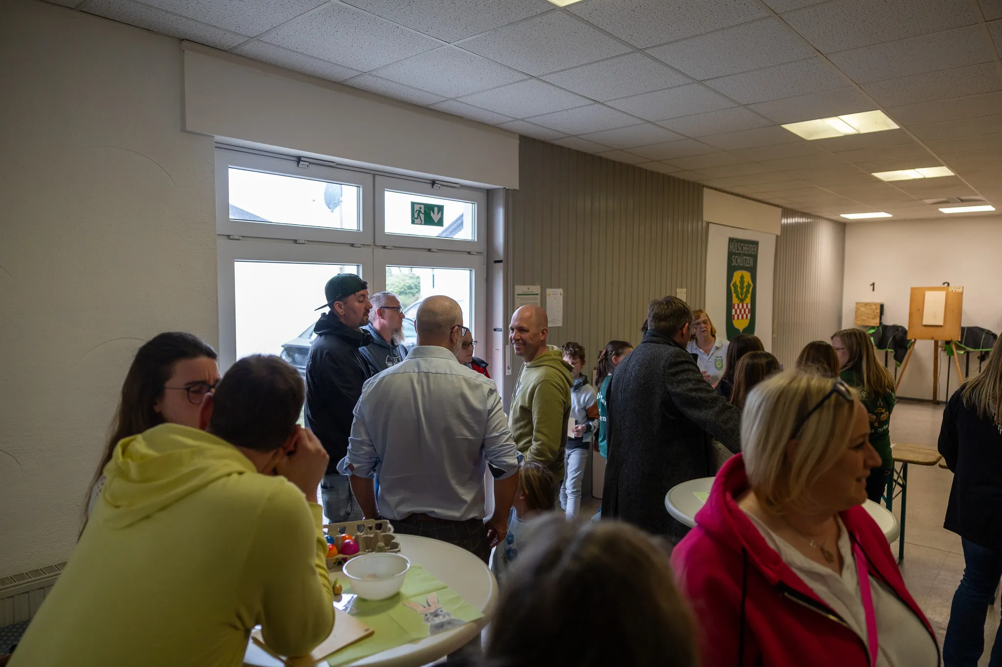 Menschen stehen und unterhalten sich in einem hellen Raum, einige stehen um Tische mit Eiern. Im Hintergrund ist ein Türrahmen und ein Wappen zu sehen.