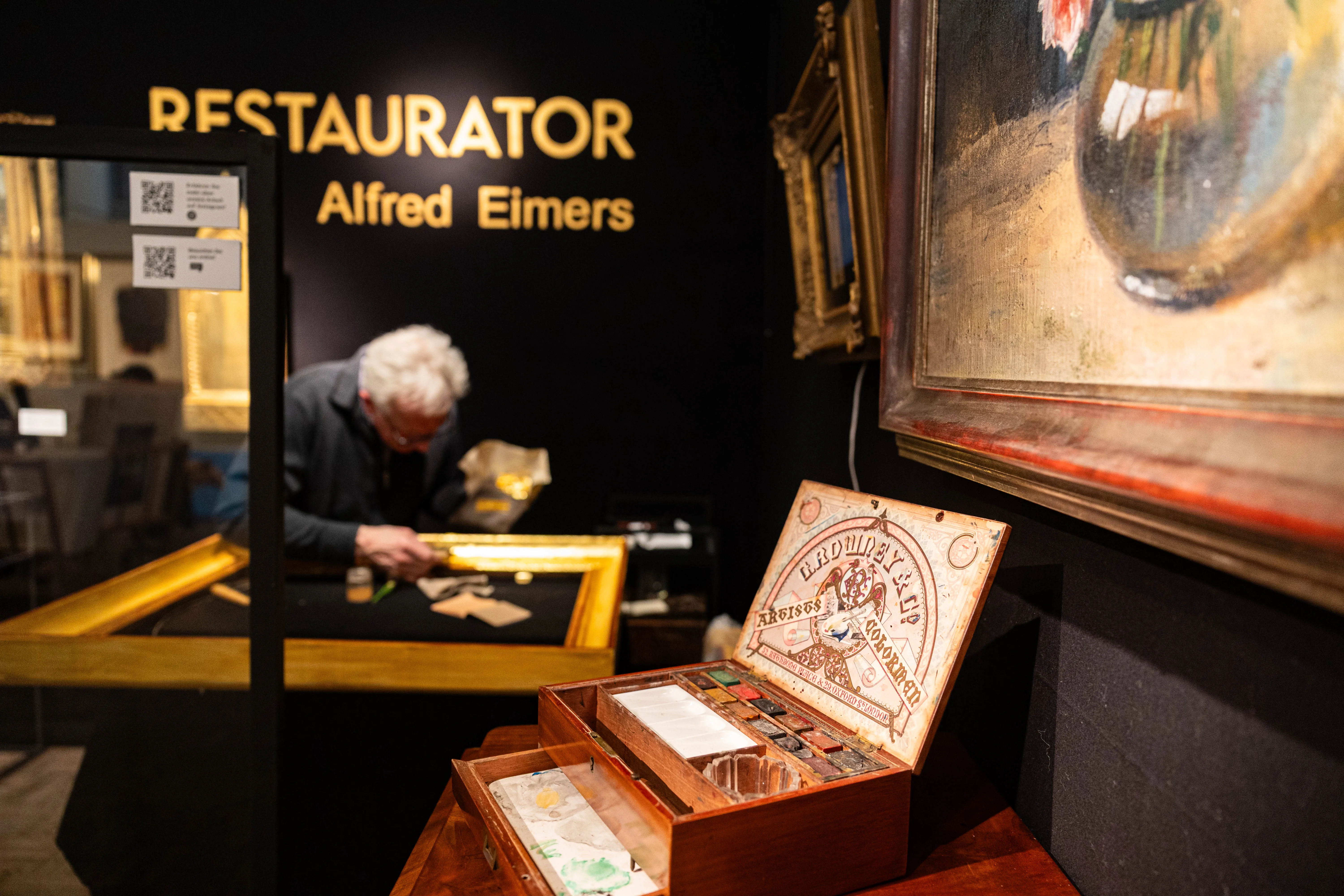 Ein Restaurator arbeitet an einem Gemälde, während im Hintergrund ein weiteres Bild an der Wand hängt. Im Vordergrund steht eine Werkzeugkiste mit Pinseln und Farben.