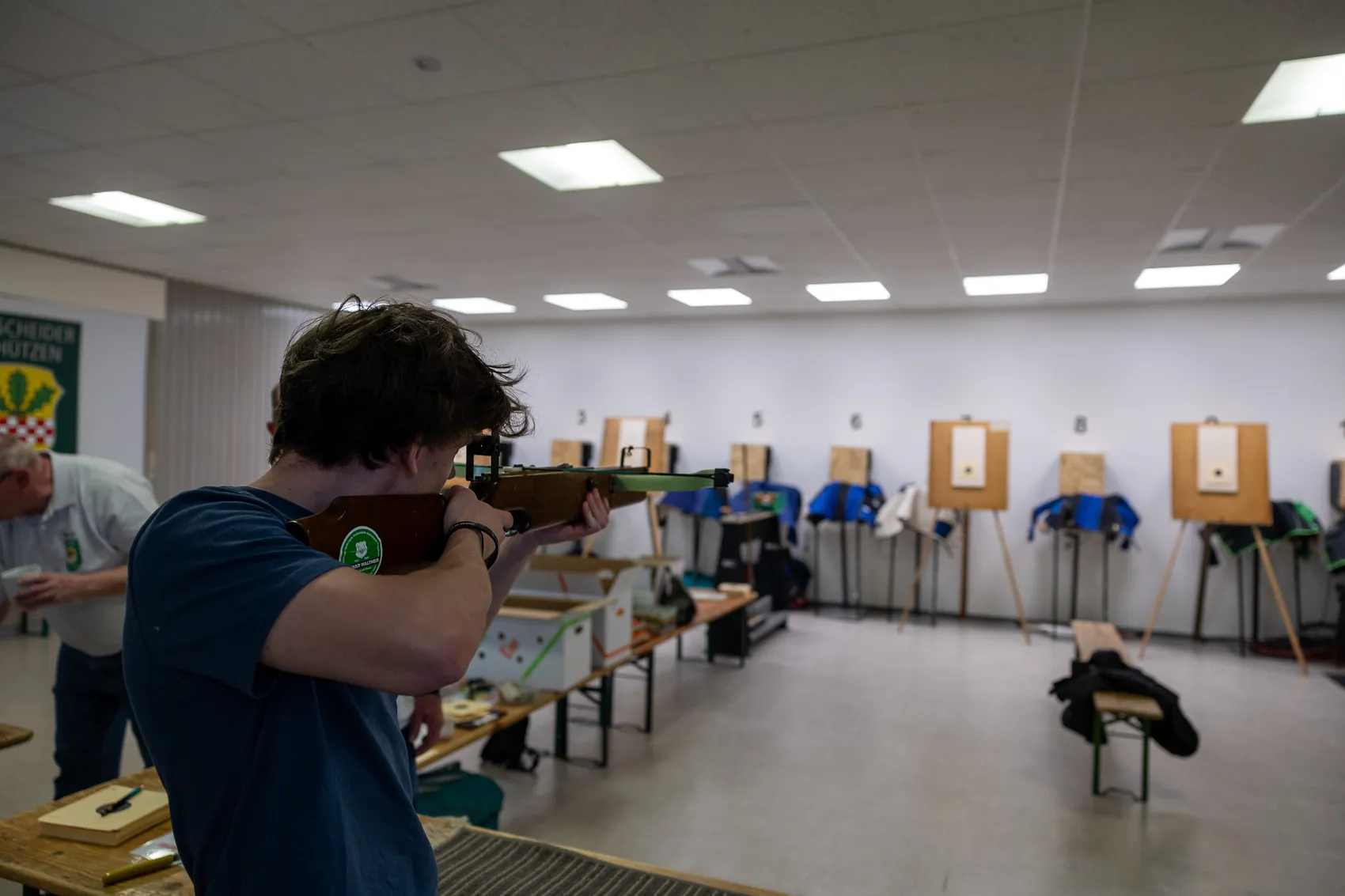 Ein junger Mann zielt mit einem Gewehr in einer Schießhalle auf eine Zielscheibe. Im Hintergrund sind weitere Schützen und Zielscheiben zu sehen.