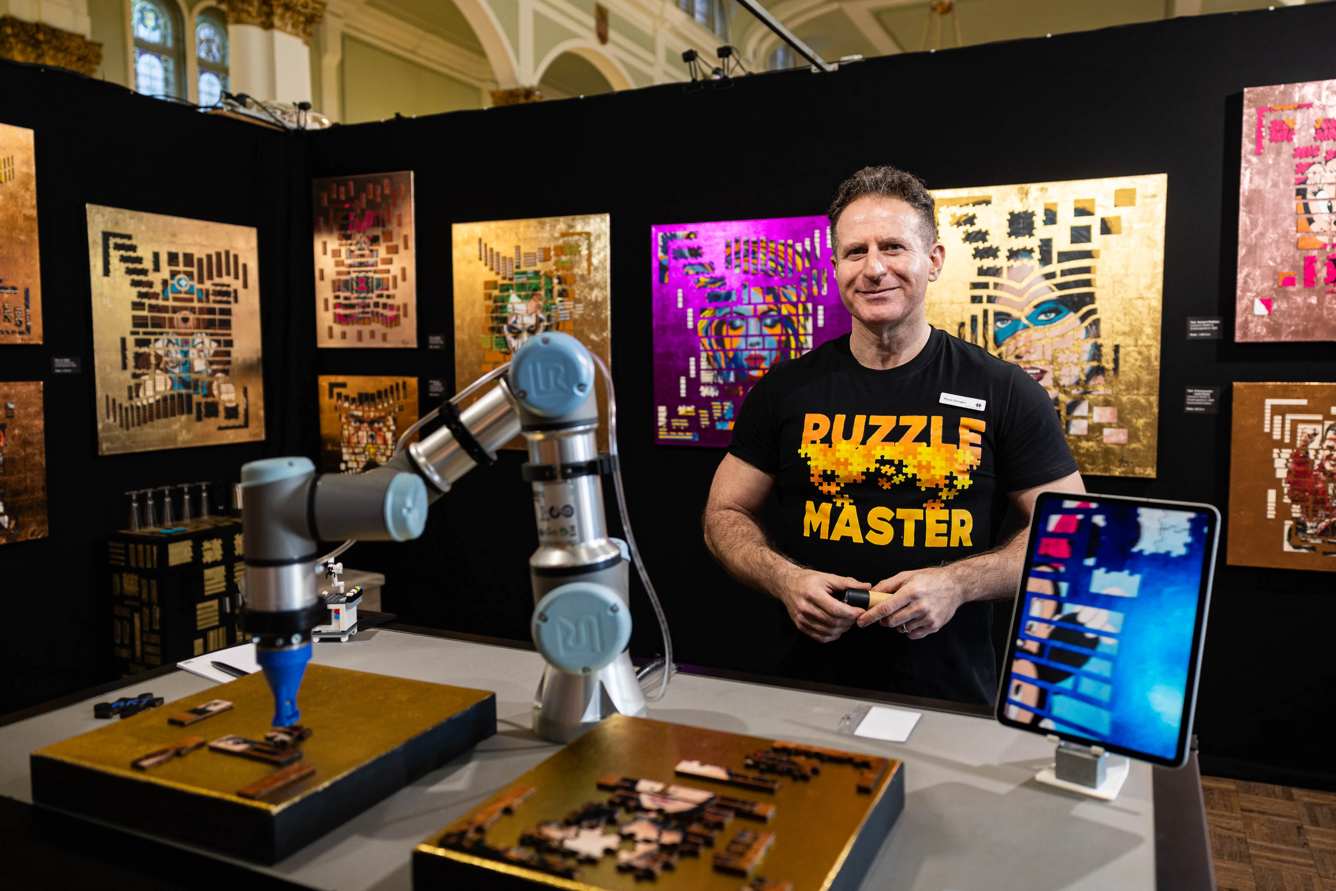 Ein Mann mit T-Shirtaufdruck 'Puzzle Master' steht vor einer Ausstellung von farbenfrohen, abstrakten Kunstwerken und einem Roboterarm, der Puzzleteile zusammensetzt. Im Hintergrund sind weitere Kunstwerke an einer schwarzen Wand zu sehen.