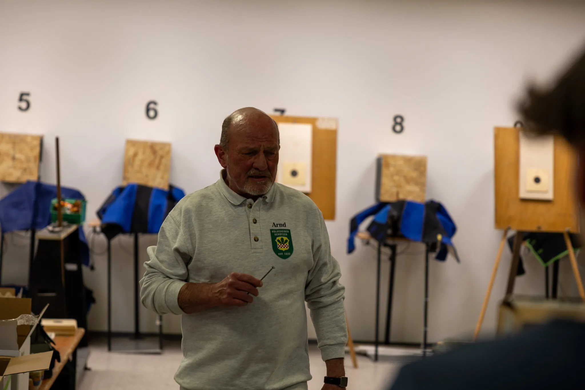 Ein älterer Mann mit Kurzhaarschnitt und grünem Pullover steht in einer Schießhalle vor mehreren Zielscheiben mit Einschusslöchern.