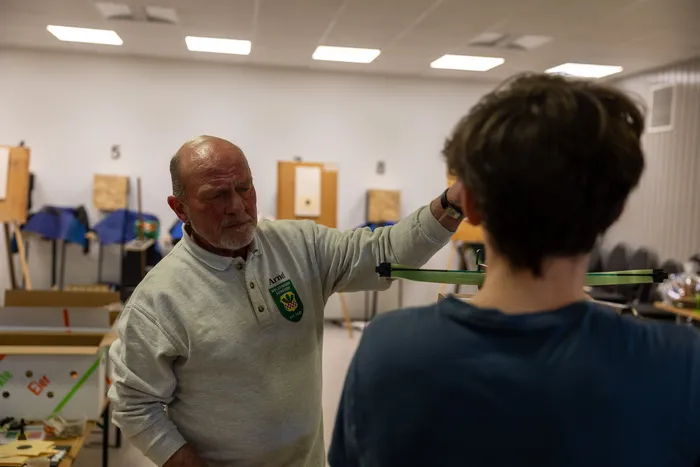 Ein älterer Mann erklärt einem Jugendlichen die Handhabung eines Bogens im Schützenverein. Im Hintergrund sind weitere Zielscheiben und Vereinsmitglieder zu sehen.