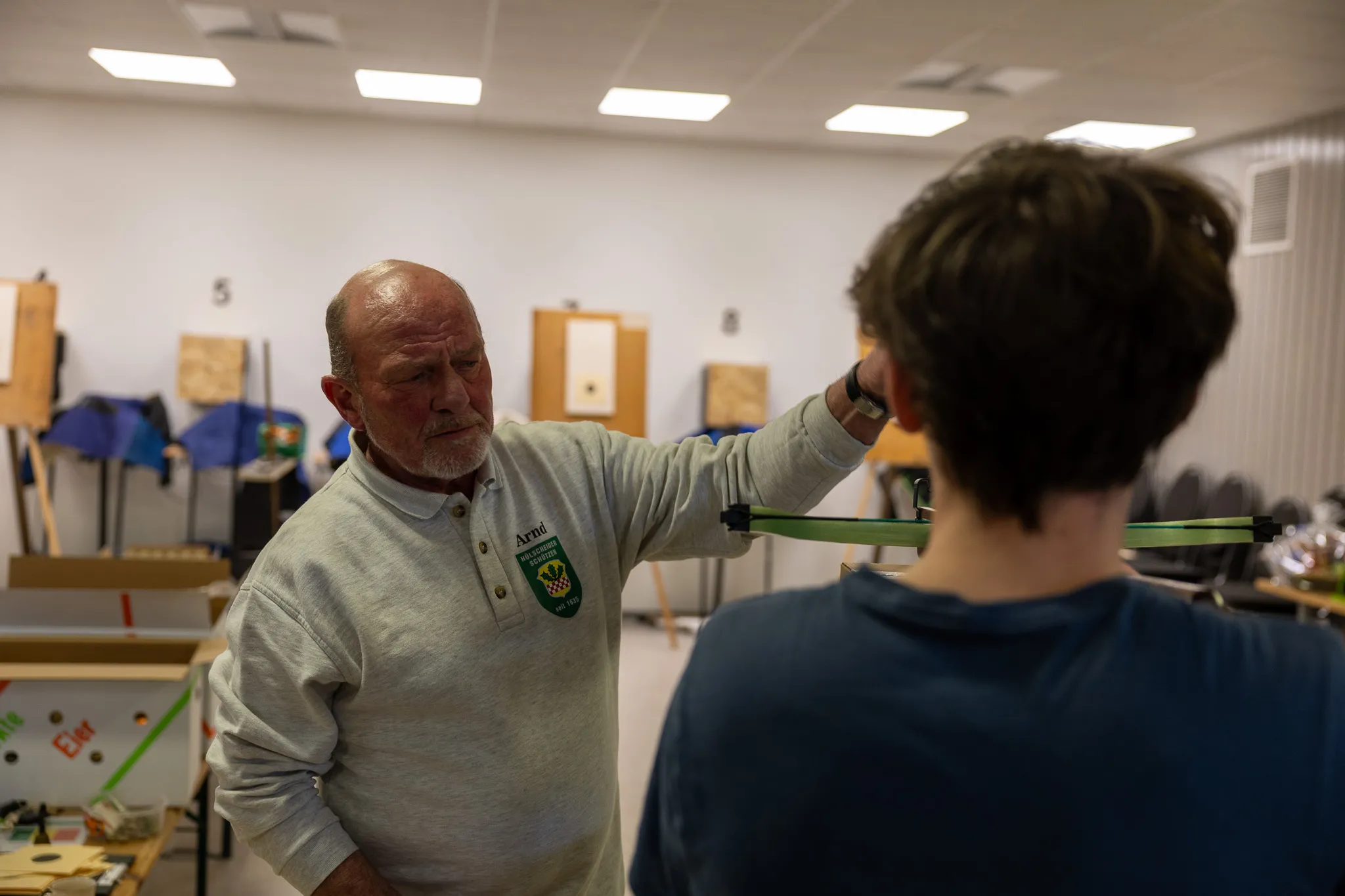 Ein älterer Mann erklärt einem Jugendlichen die Handhabung eines Bogens im Schützenverein. Im Hintergrund sind weitere Zielscheiben und Vereinsmitglieder zu sehen.