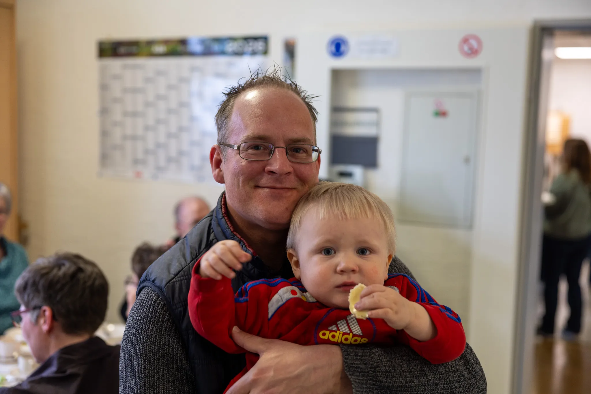 Ein Mann hält ein Kleinkind im Arm, das in die Kamera schaut und etwas isst. Im Hintergrund sind weitere Personen und eine Tür zu sehen.