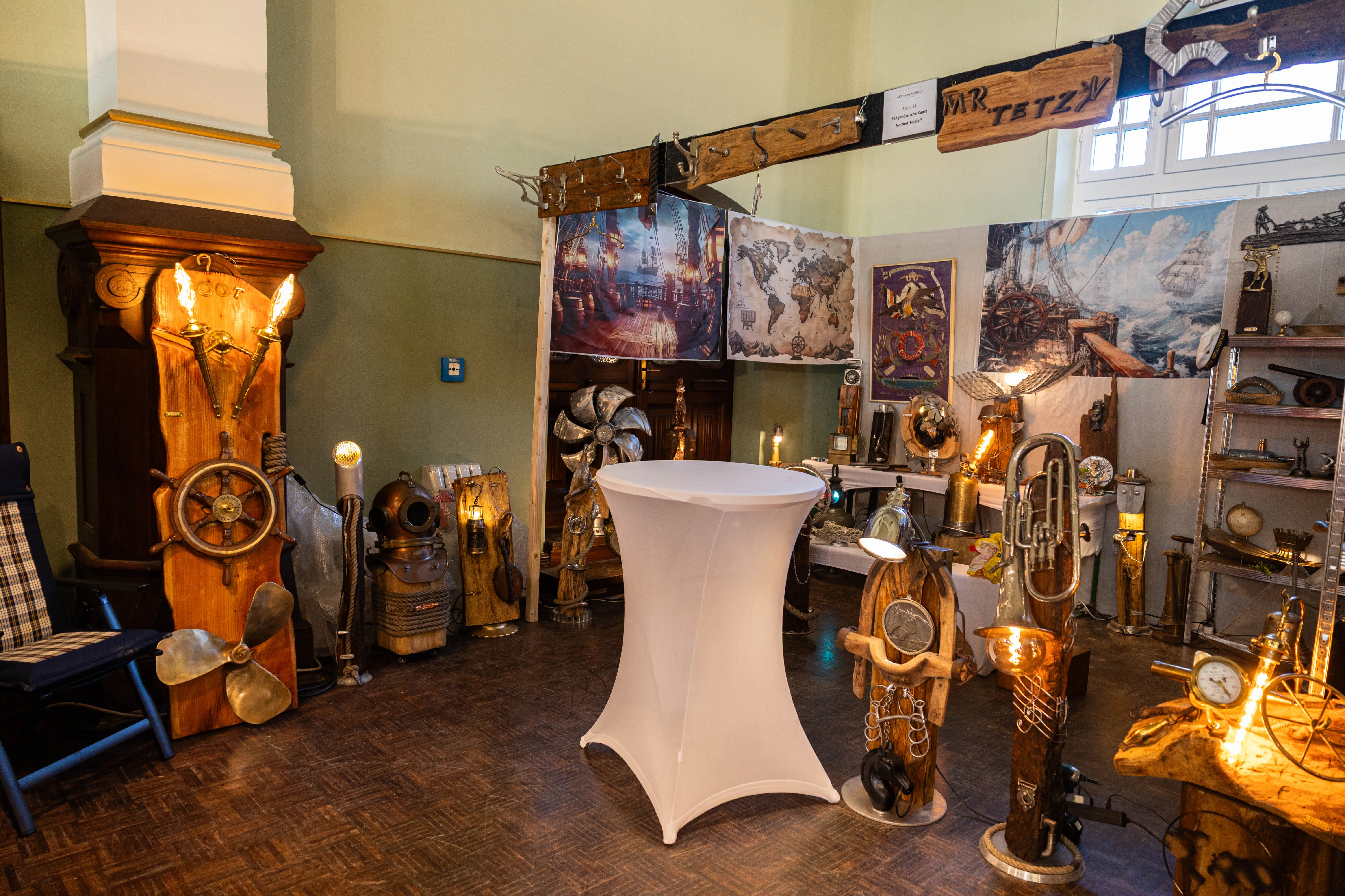Ausstellungsraum mit antiken und modernen Kunstobjekten, darunter Möbel, Lampen und Gemälde mit maritimen Motiven. Im Zentrum steht ein runder, weiß bezogener Tisch.