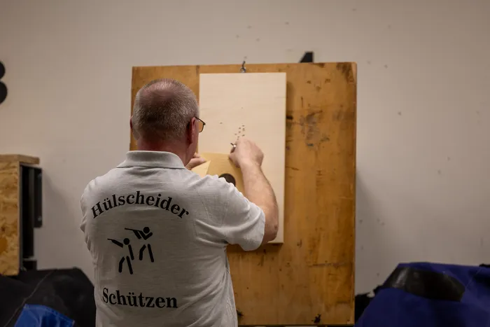 Ein Mann mit grauem Haar und einem hellgrauen Pullover mit dem Aufdruck „Hülscheider Schützen“ zielt mit einem Gewehr auf eine Zielscheibe.