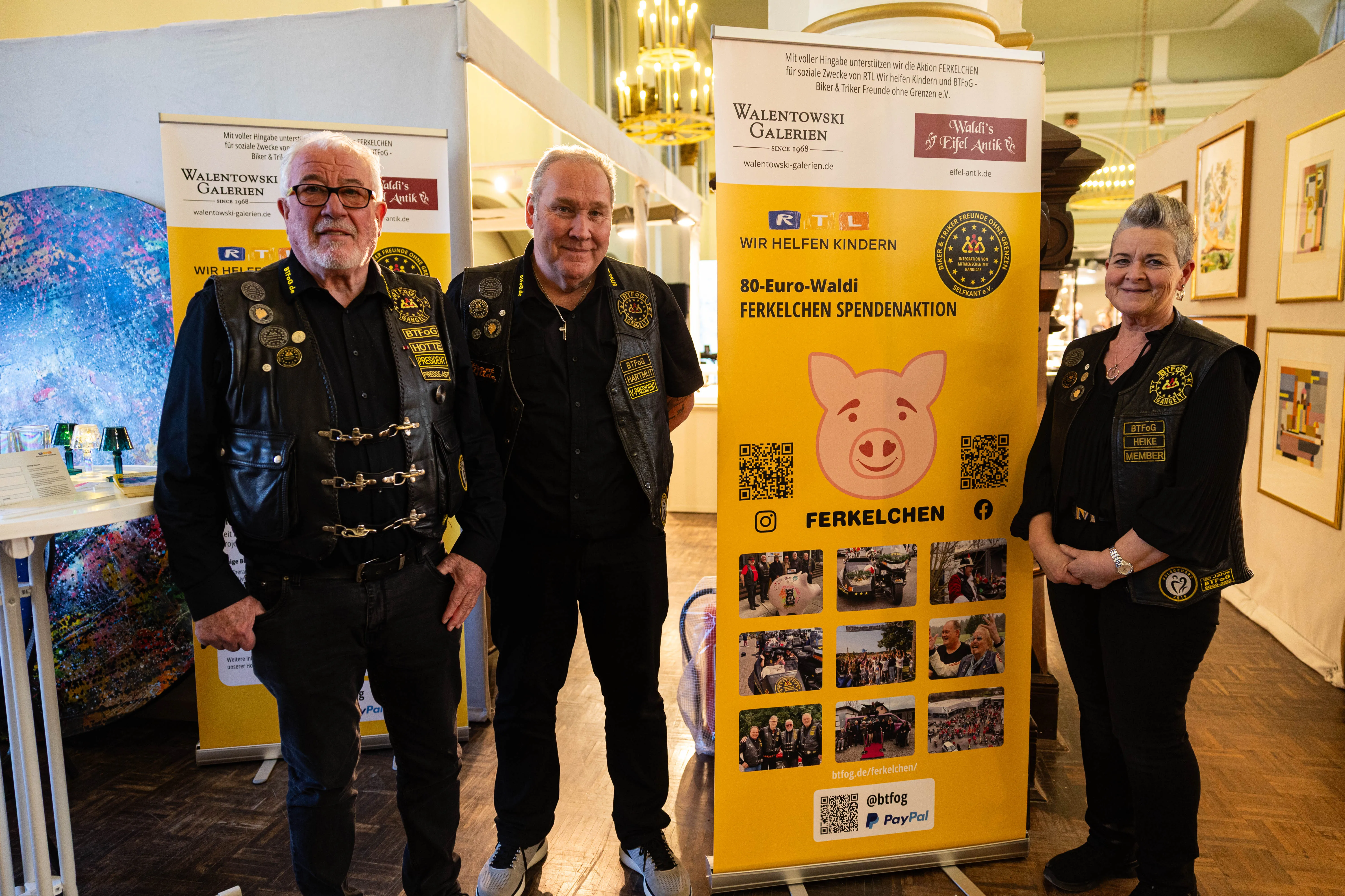 Vier Personen stehen vor einem Roll-up-Banner mit dem Aufdruck „80-Euro-Waldi – Ferkelchen Spendenaktion“. Die drei Männer und eine Frau tragen Lederwesten mit Aufnähern und lächeln in die Kamera.