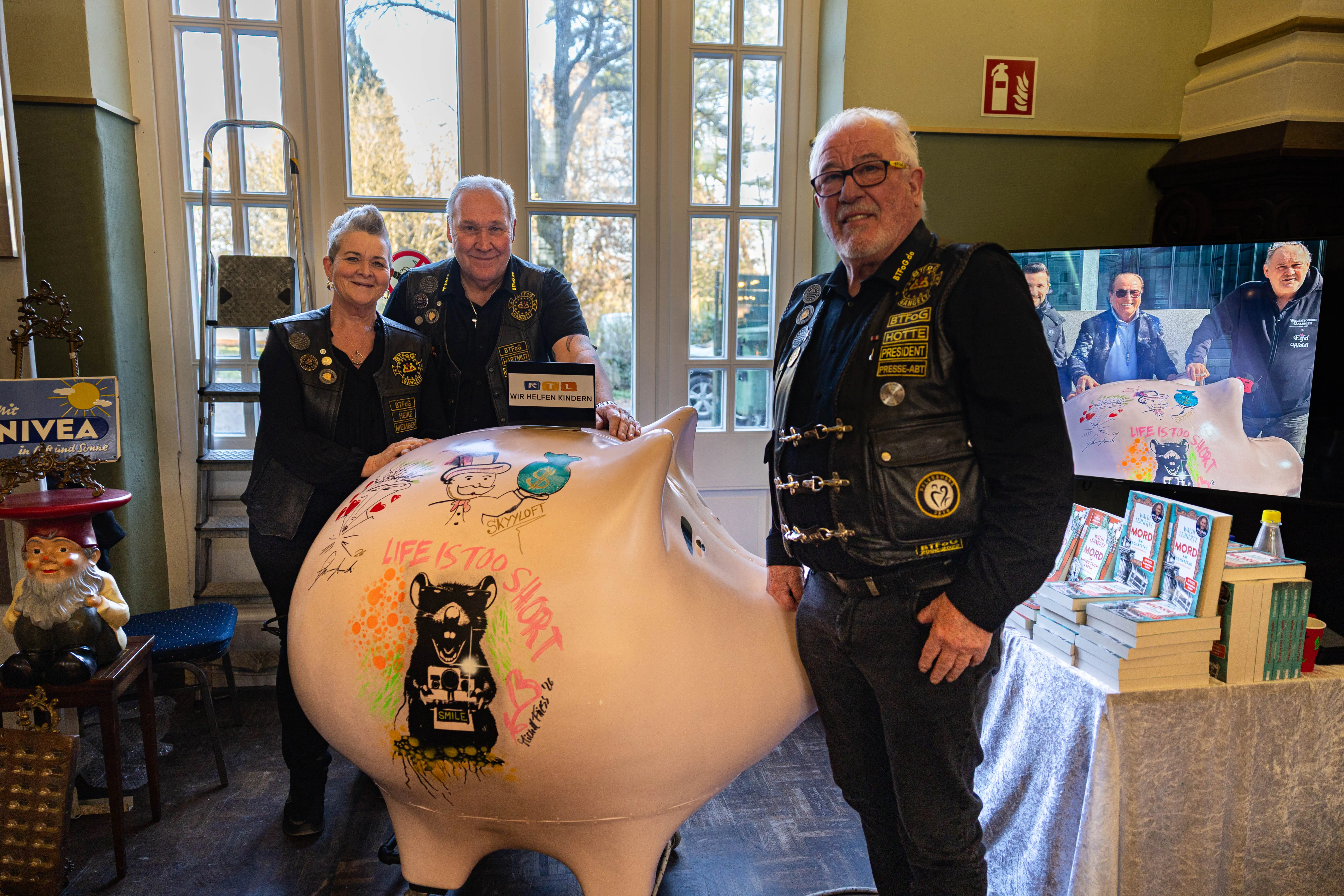 Zwei Motorradfahrer und eine Motorradfahrerin stehen neben einem großen, bemalten Sparschwein mit der Aufschrift „Life is too short“. Im Hintergrund ist ein Bildschirm mit weiteren Personen zu sehen.