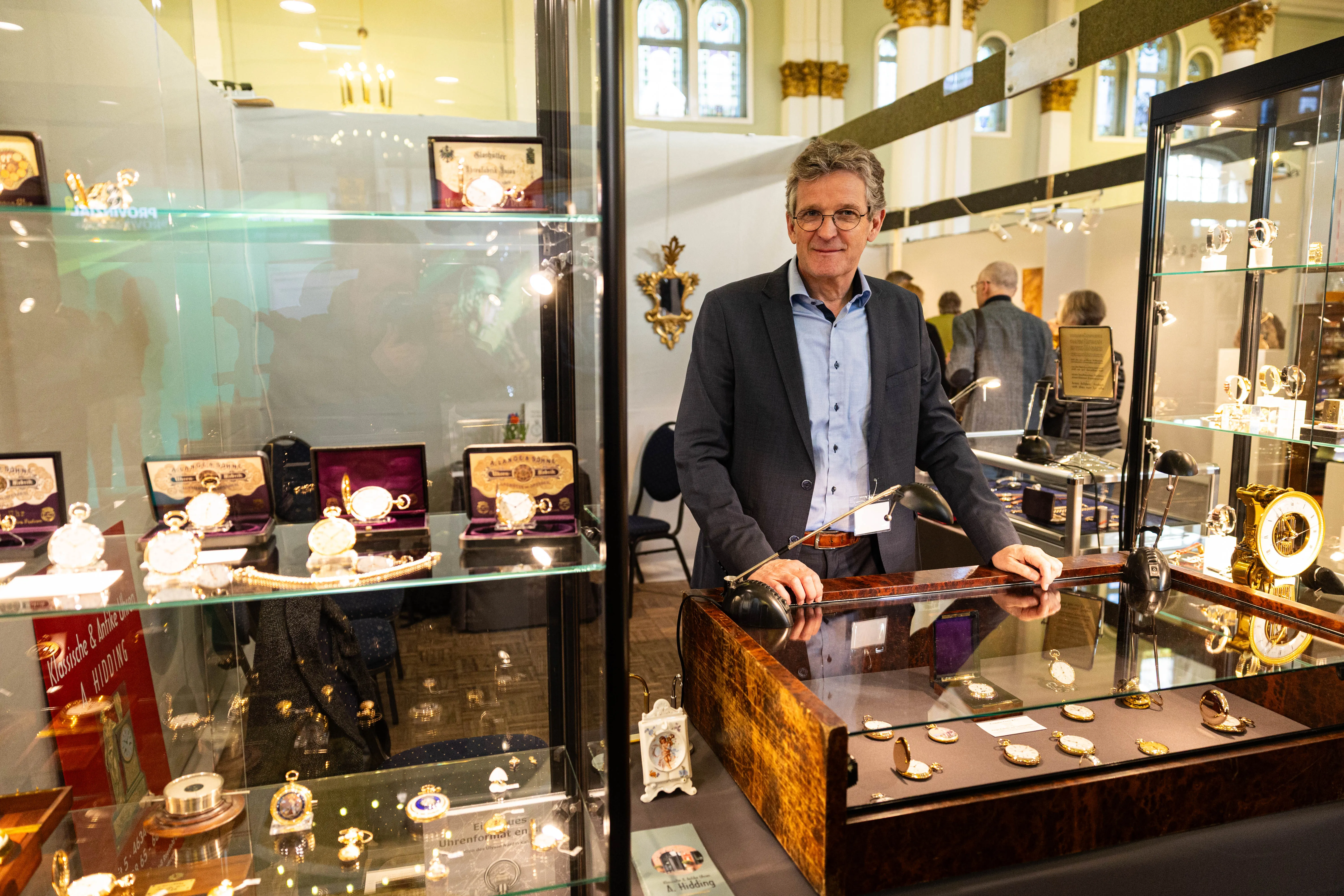 Ein Mann mit Brille und Anzug steht lächelnd vor einer Vitrine mit antiken Taschenuhren und Schmuck auf einer Messe.