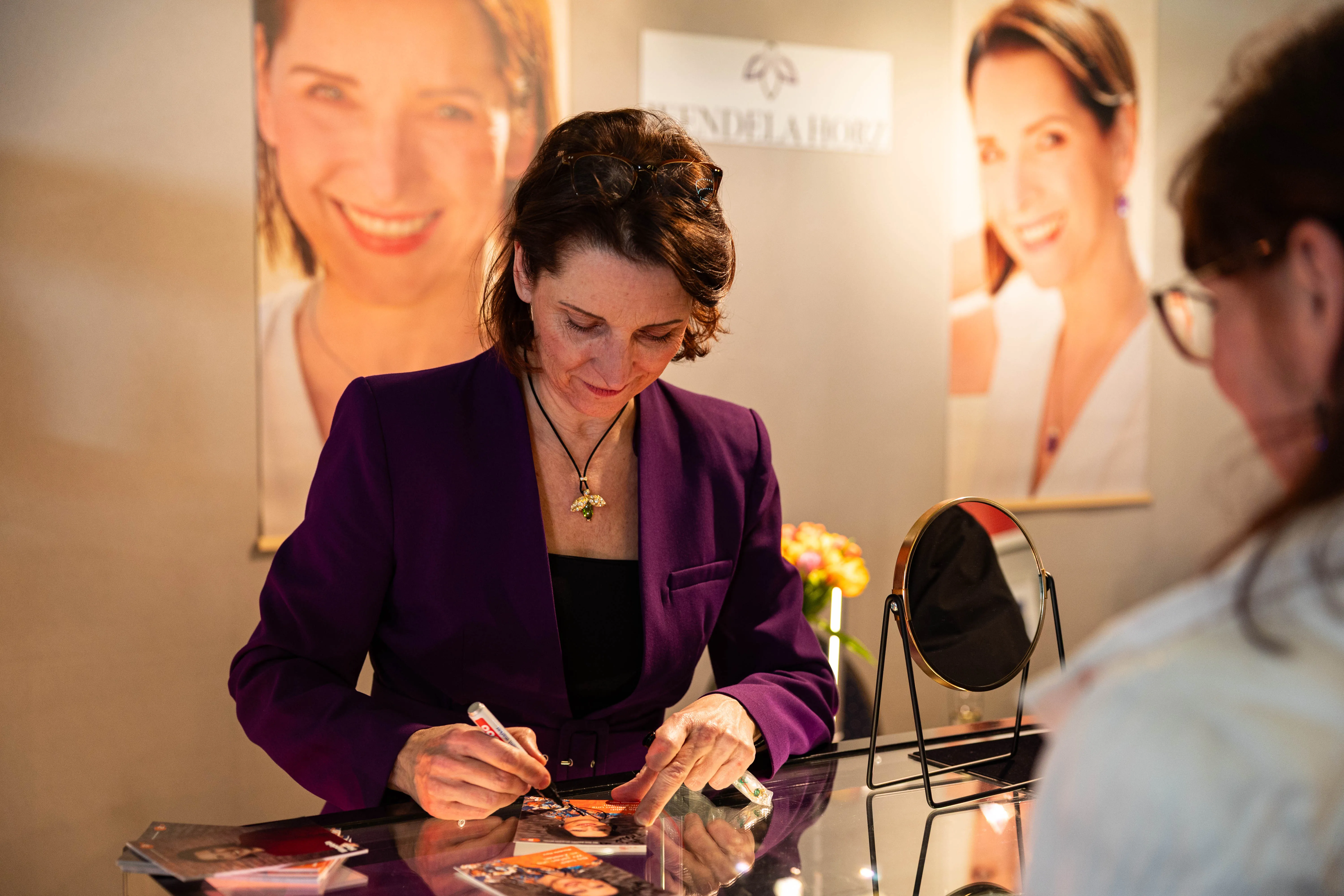 Wendela Horz signiert auf einer Vernissage in Lüdenscheid Autogrammkarten. Im Hintergrund sind Porträtbilder und ein Blumenstrauß zu sehen.