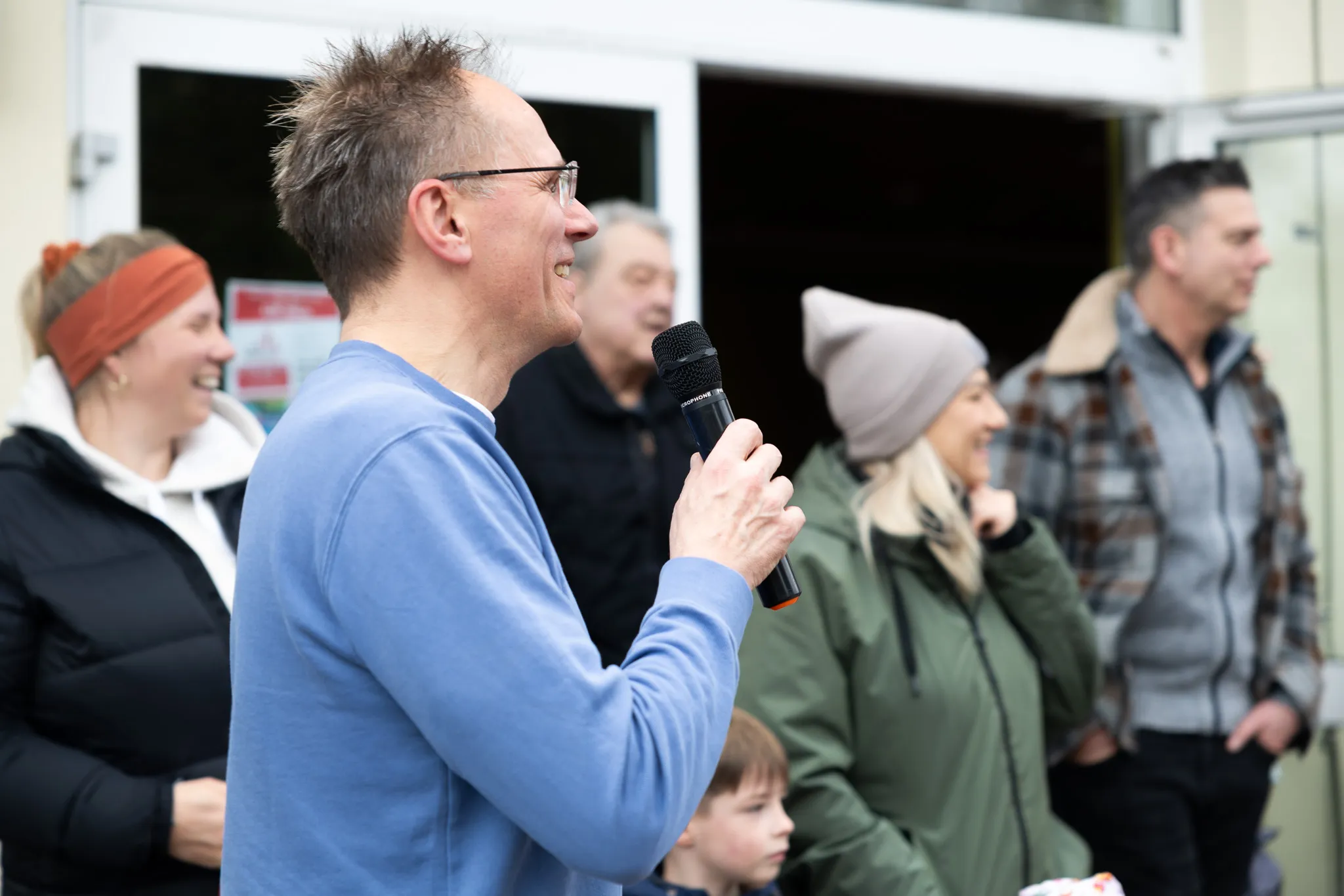 Ein Mann mit Brille spricht in ein Mikrofon, umgeben von Familien bei einer Ostereiersuche im Freien.