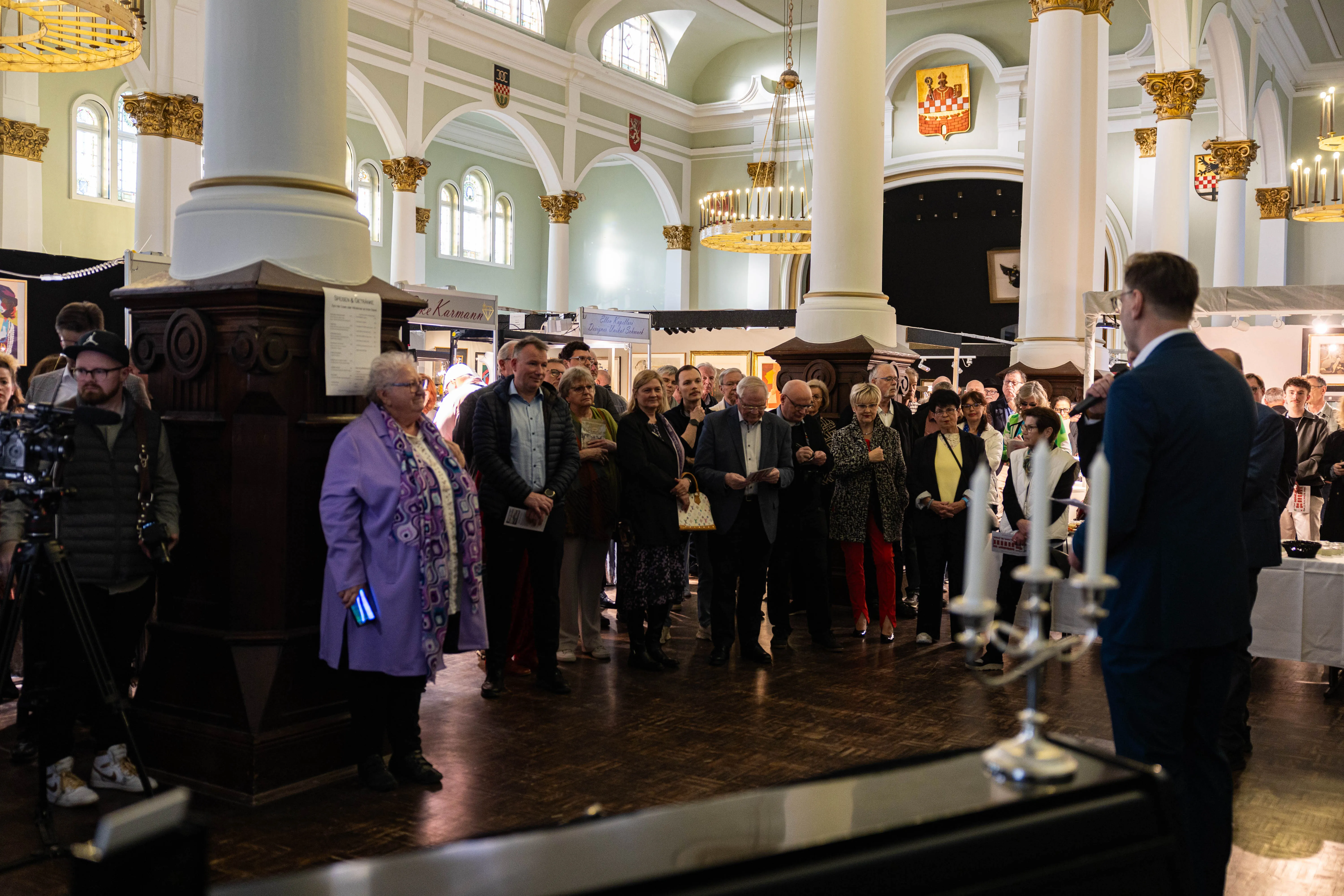 Eine Gruppe von Menschen steht in einer hellen Kirche und betrachtet Kunstwerke. Im Vordergrund steht ein Mann in einem lilafarbenen Gewand.