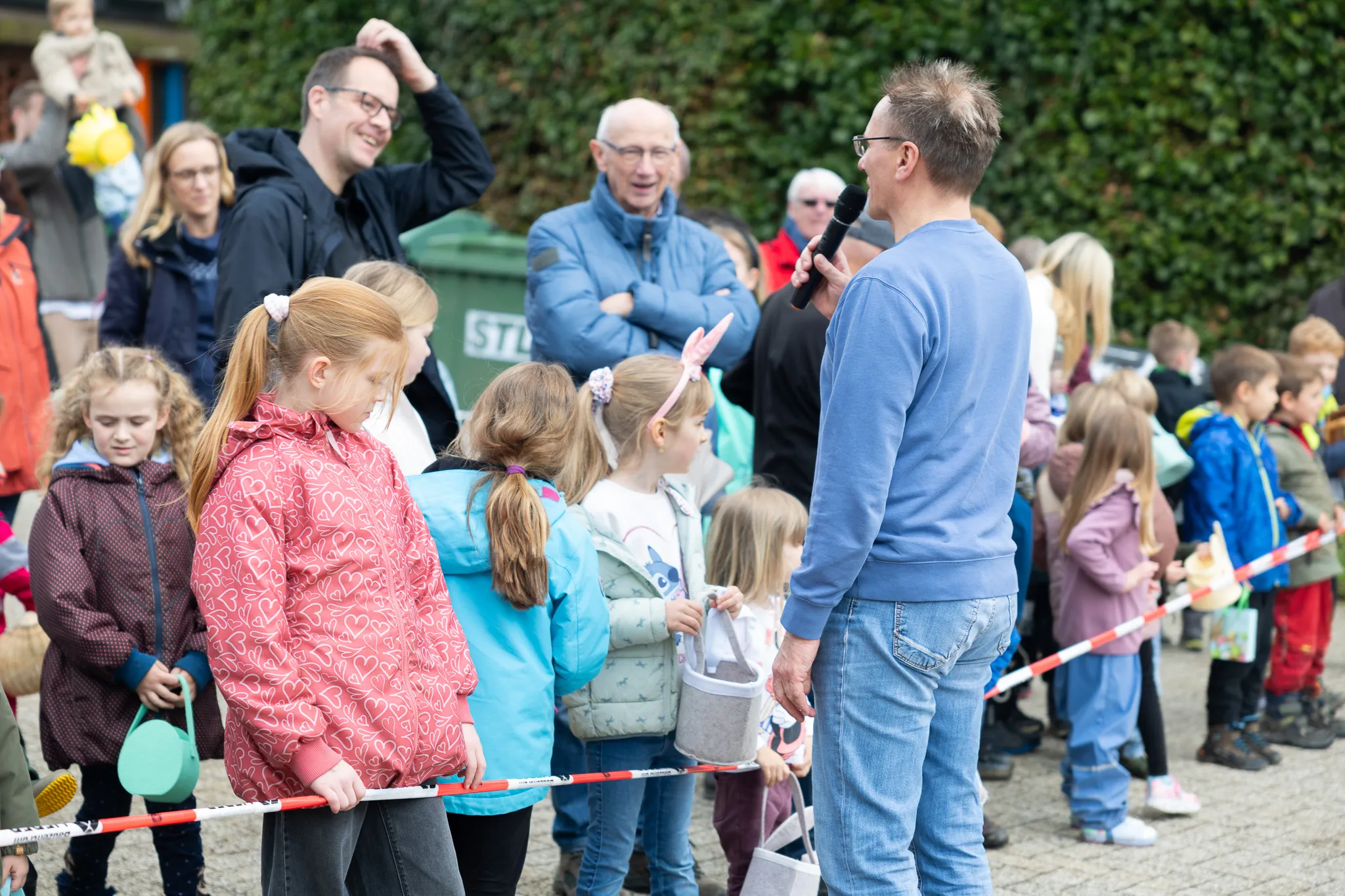 Ein Mann spricht in ein Mikrofon zu einer Gruppe Kinder bei einer Ostereiersuche, umgeben von Eltern und Zuschauern.