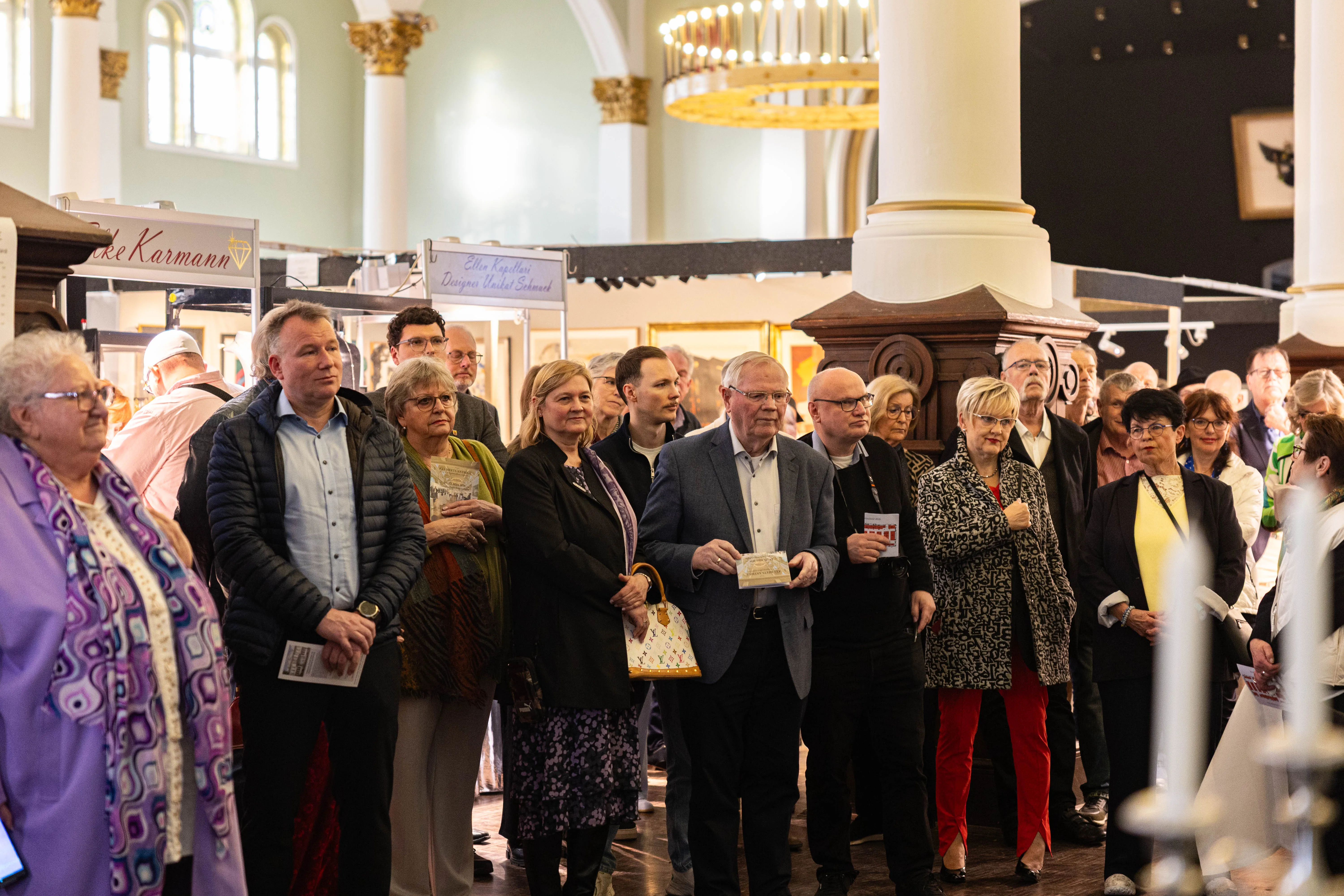 Besucher stehen vor Kunstwerken in einer hellen, hohen Halle mit Kirchenfenstern. Im Vordergrund hält ein Mann in einem grauen Anzug ein kleines Geschenk in den Händen.