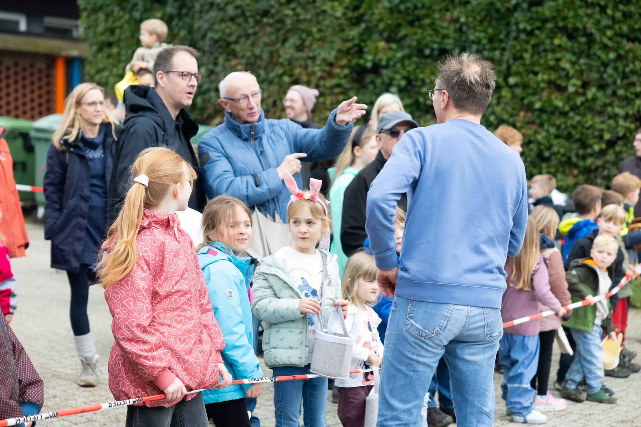 Eine Gruppe Kinder mit Ostereiern steht vor einem Mann, der etwas erklärt. Im Hintergrund stehen weitere Familien und Zuschauer bei der Ostereiersuche.