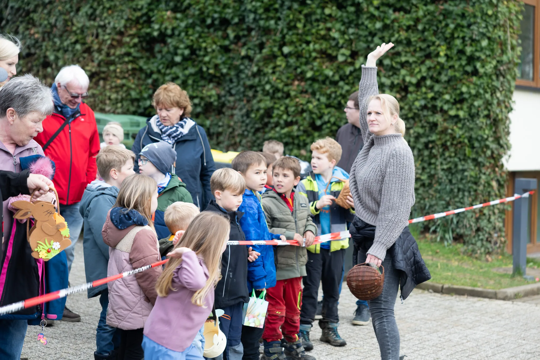 Eine Frau mit hochgehaltenem Arm weist eine Gruppe Kinder und ältere Begleitpersonen bei einer Ostereiersuche aus. Die Szene spielt vor einer Hecke und einem Gebäude mit roter Markierung.