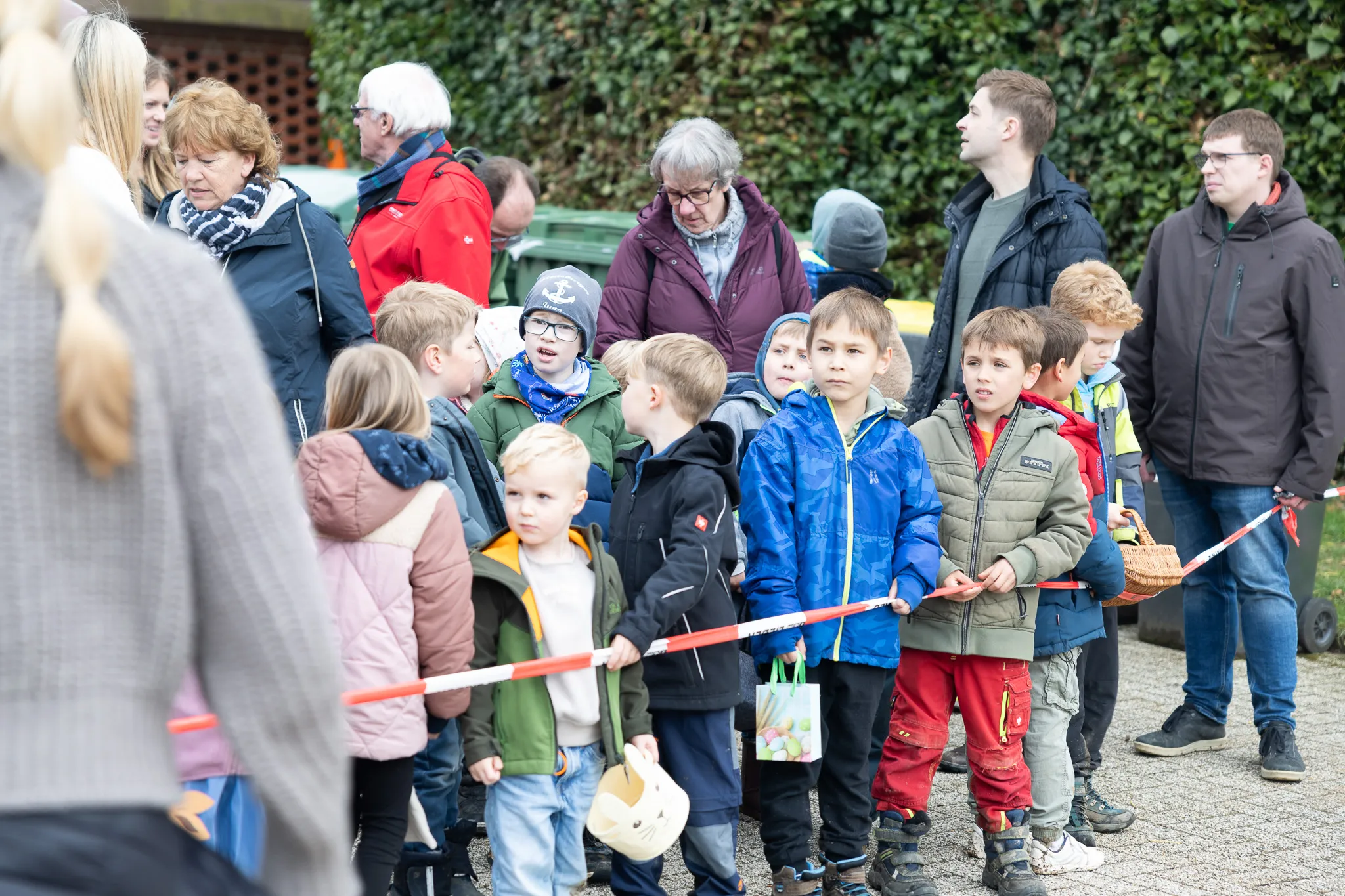 Eine Gruppe Kinder steht hinter einer roten Absperrung, viele halten Ostereierkörbe bereit. Im Hintergrund stehen Erwachsene und beobachten die Szene.