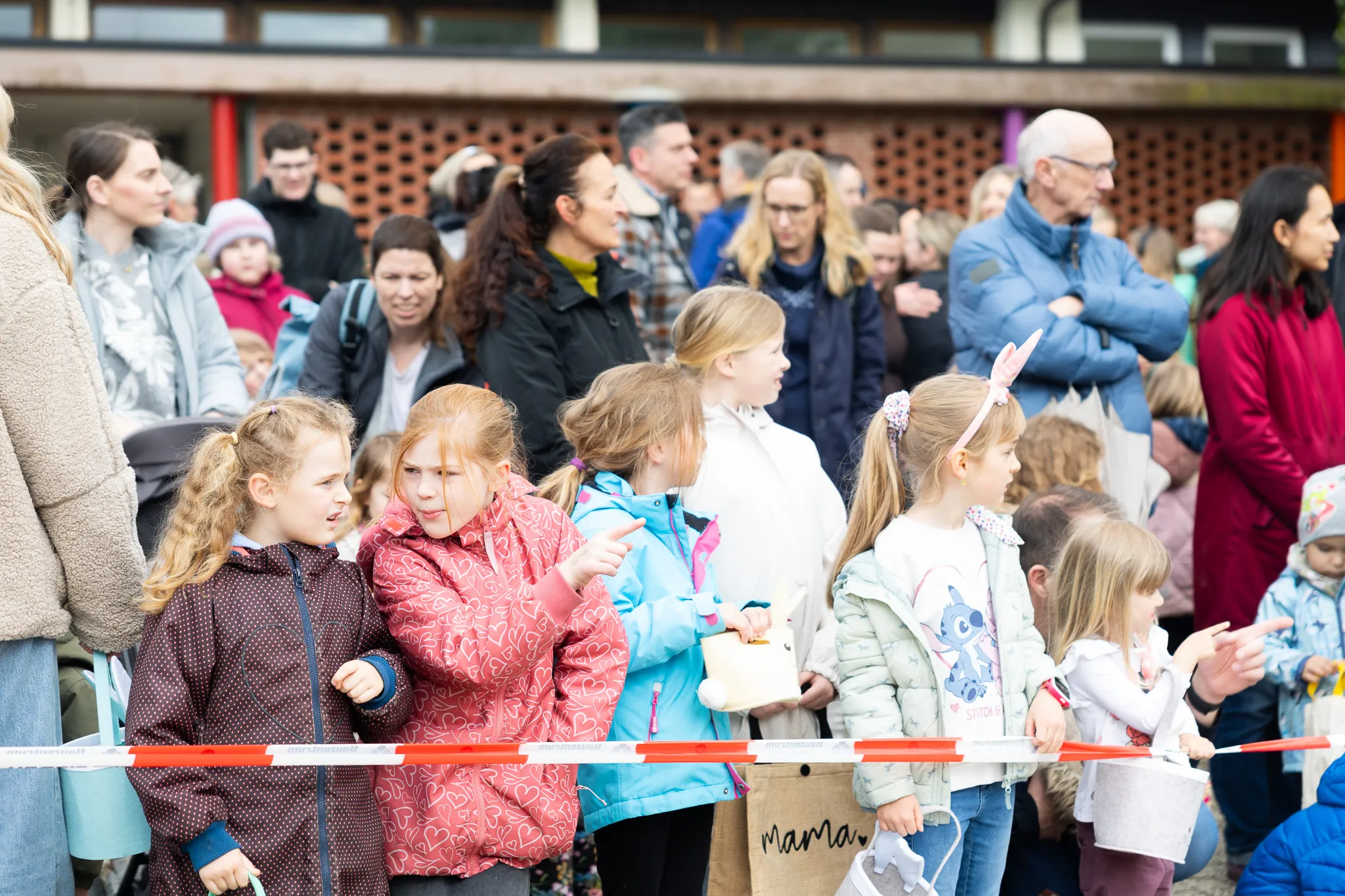 Mehrere Kinder und Eltern stehen hinter einer roten Absperrung und schauen gespannt nach vorne, vermutlich auf eine Ostereiersuche.