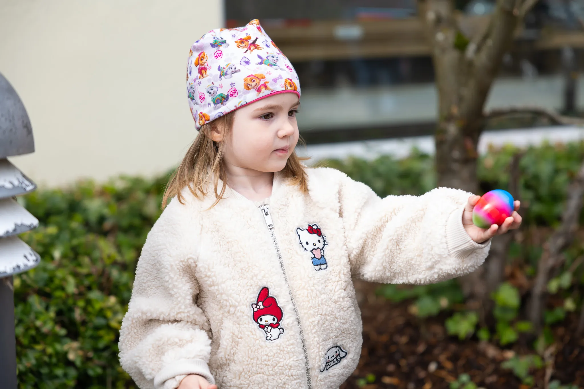 Kleines Mädchen mit bunter Mütze und Teddyfelljacke hält ein buntes Osterei in der Hand.