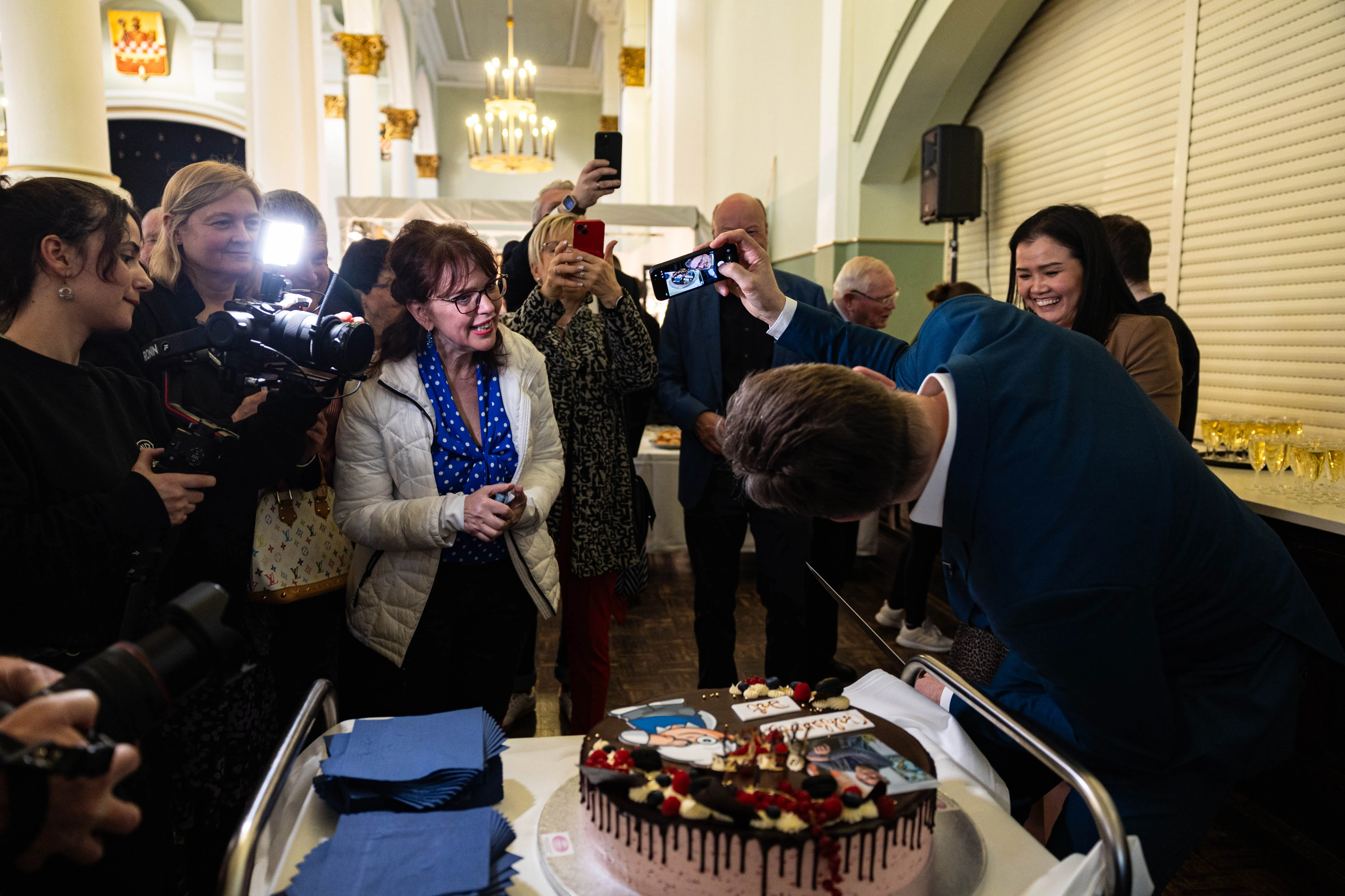 Detlev Kümmel schneidet mit einem Messer eine Geburtstagstorte vor einer Gruppe von Gästen in einem hellen Raum. Im Hintergrund fotografieren Menschen mit ihren Mobiltelefonen.