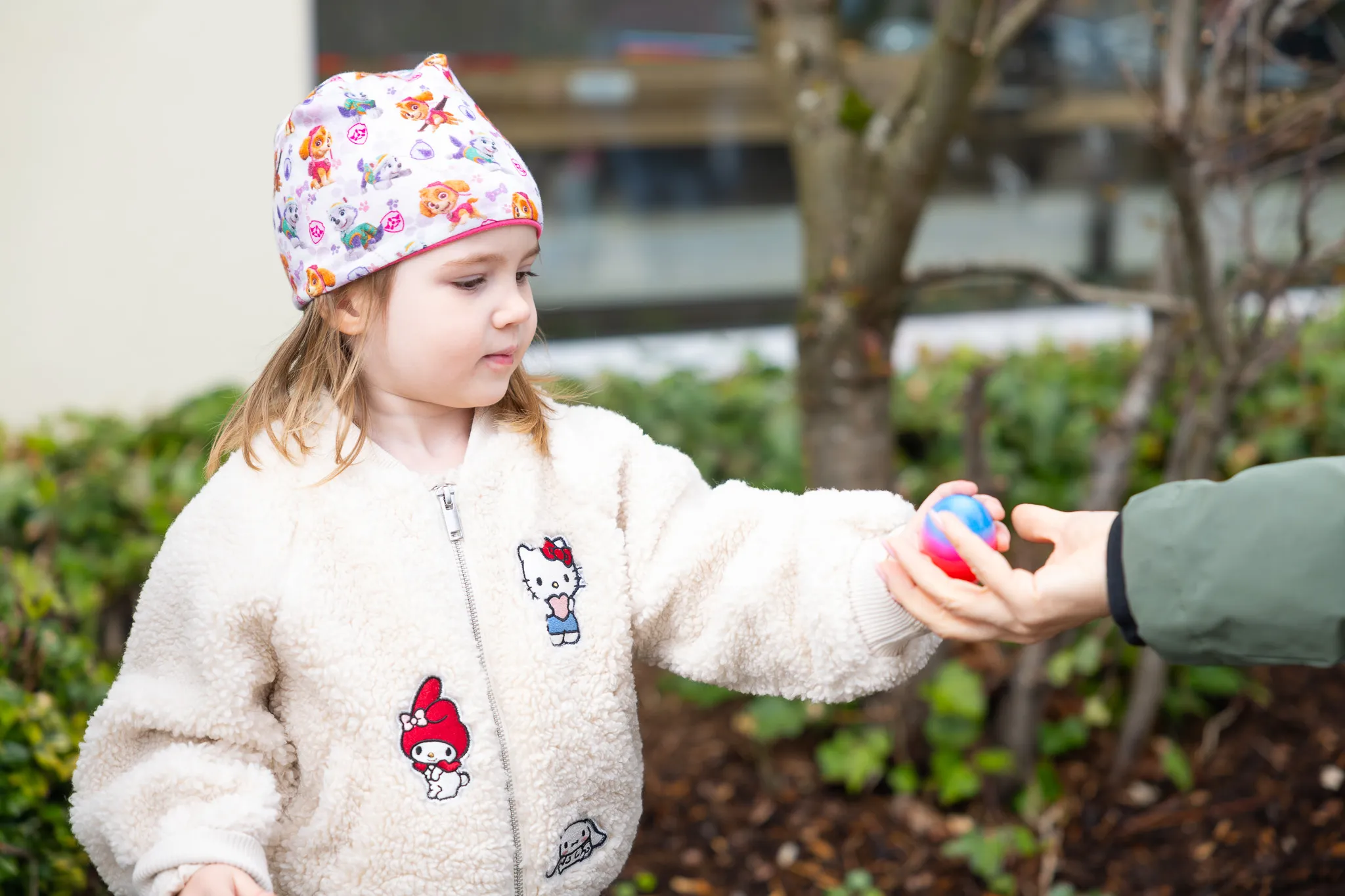 Ein kleines Mädchen in einem gemusterten Stirnband und einem Teddyfelljackett nimmt ein Osterei von einer Hand entgegen.