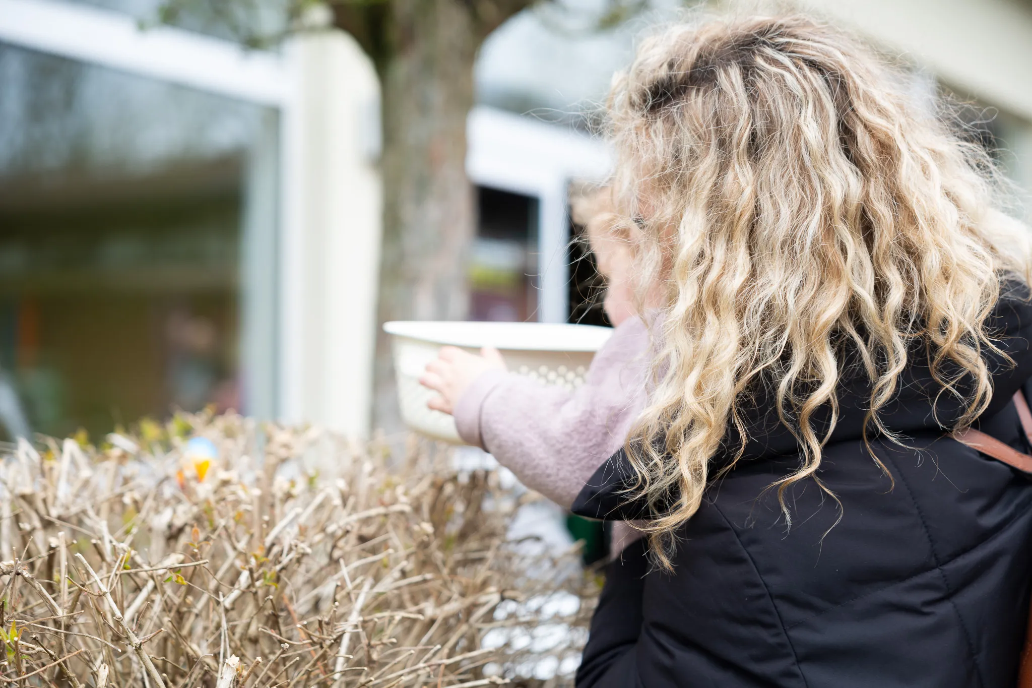 Eine Person mit gelocktem blondem Haar reicht über eine Hecke eine Schale mit Ostereiern.