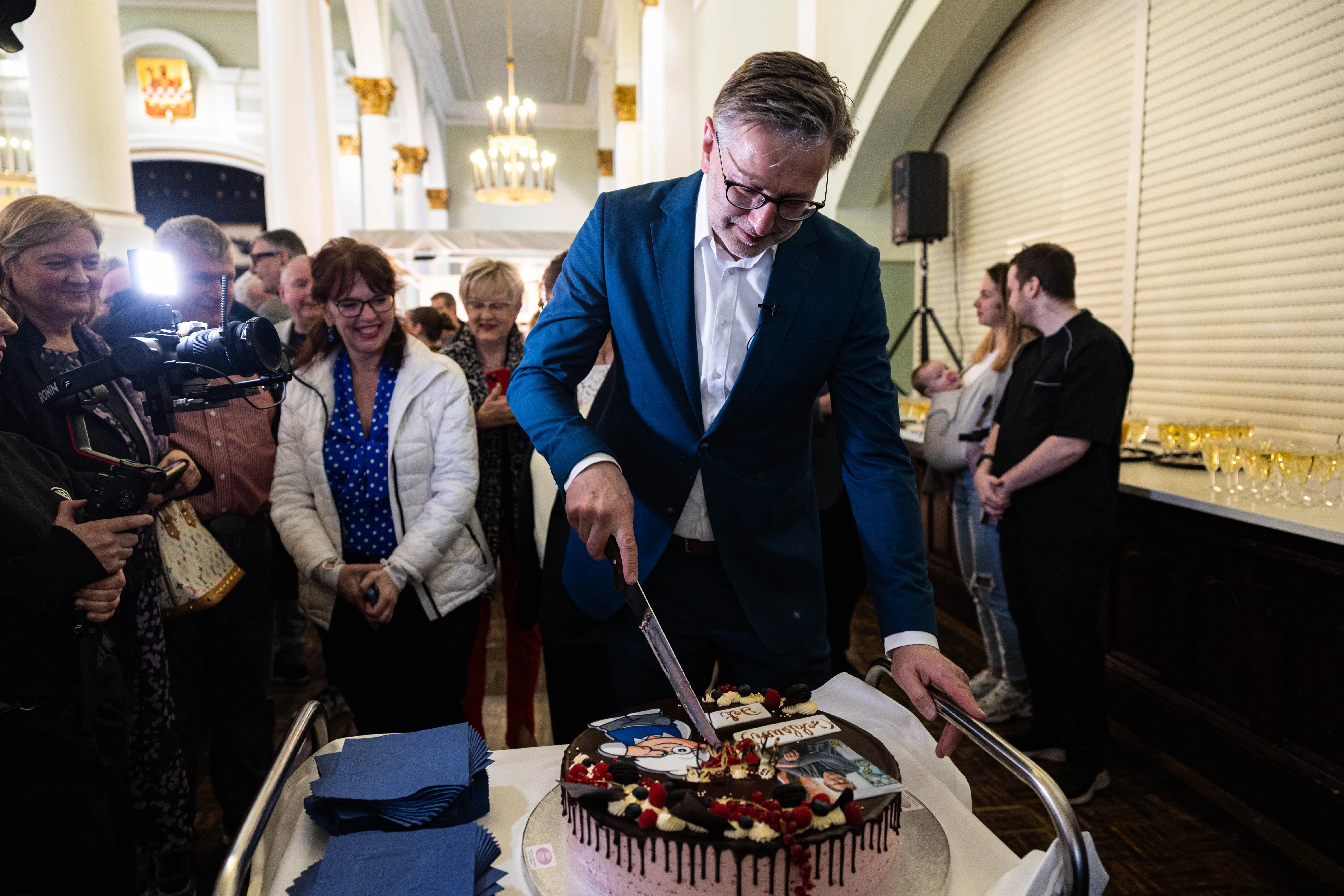 Detlev Kümmel schneidet mit einem Messer eine Geburtstagstorte vor Gästen in einem festlich dekorierten Raum.