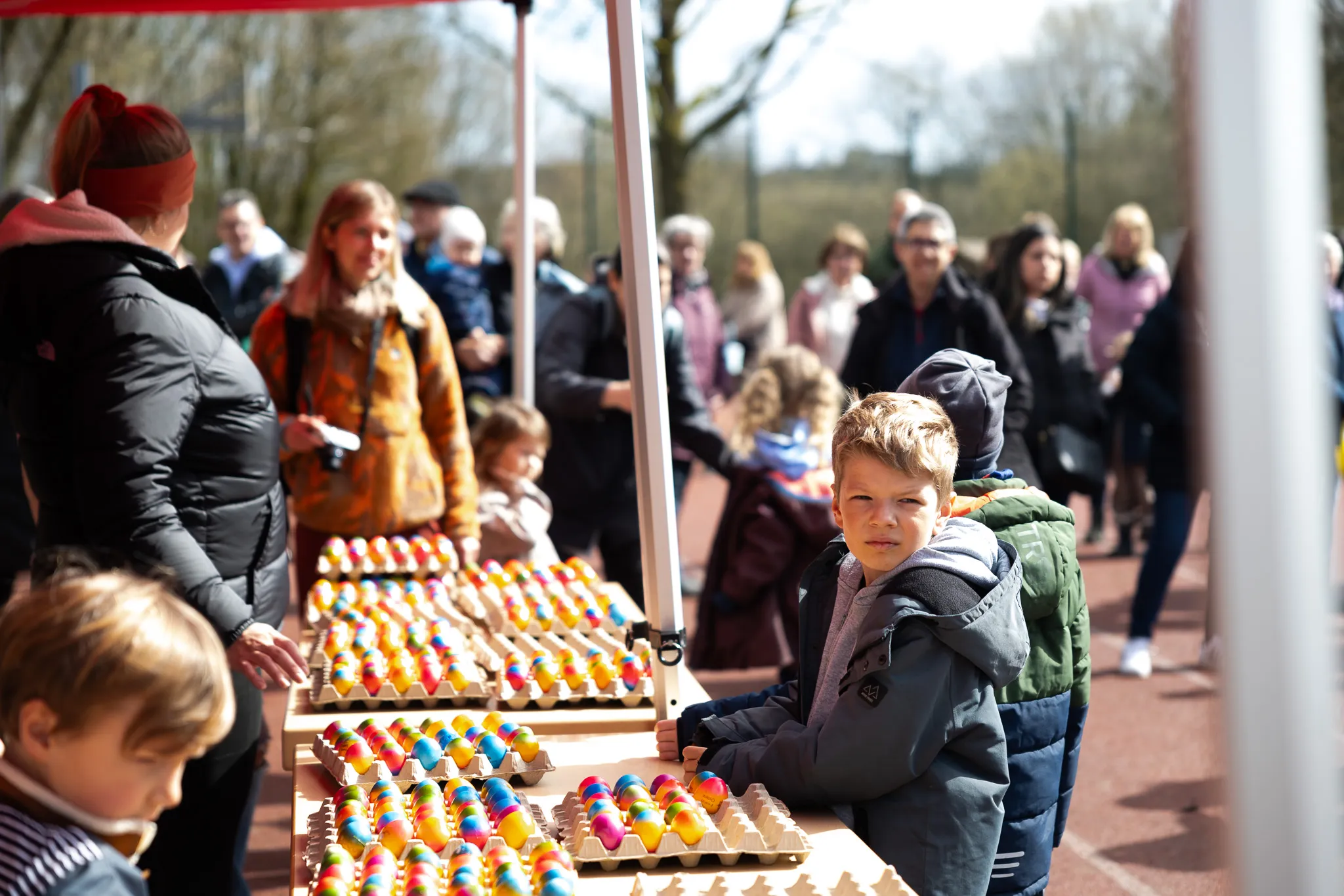 Junge steht vor einem Tisch voller bemalter Ostereier, im Hintergrund Menschen auf einem Frühlingsfest.
