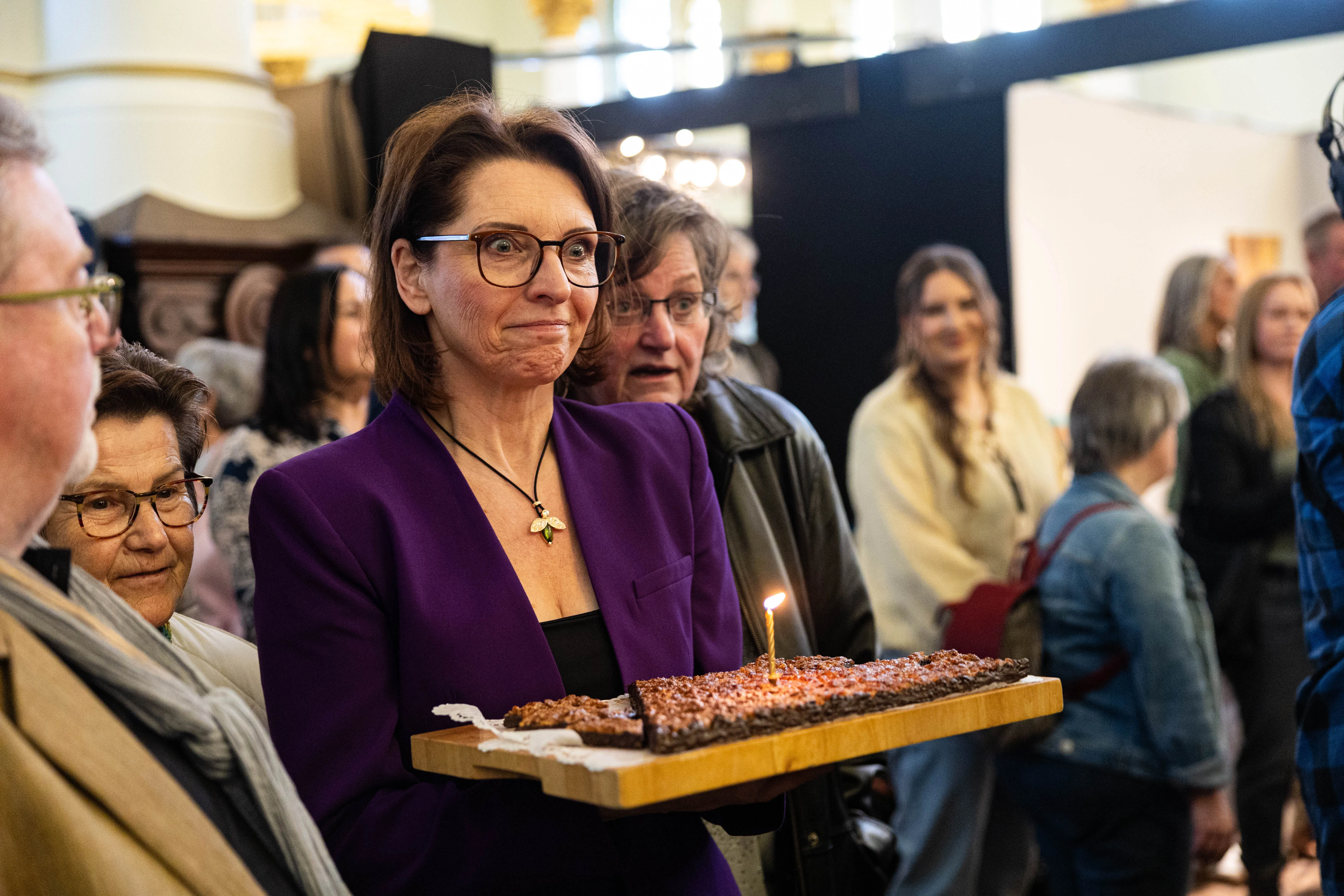 Eine Frau mit Brille und violettem Blazer trägt ein Tablett mit Nussecken, auf denen eine brennende Kerze steckt. Im Hintergrund stehen weitere Menschen in einer hellen Halle.