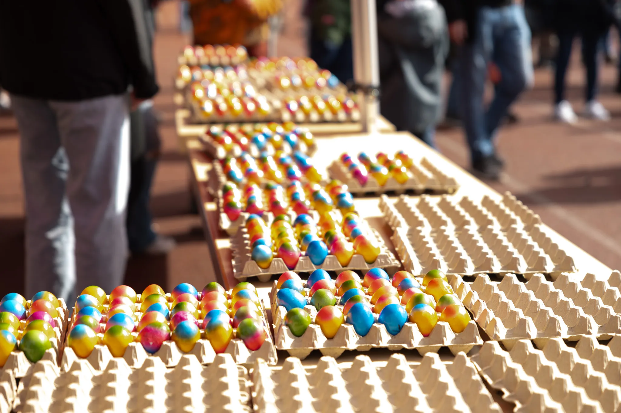 Bunte Ostereier in Pappverpackungen stehen auf einem langen Tisch im Freien, im Hintergrund verschwommene Personen.