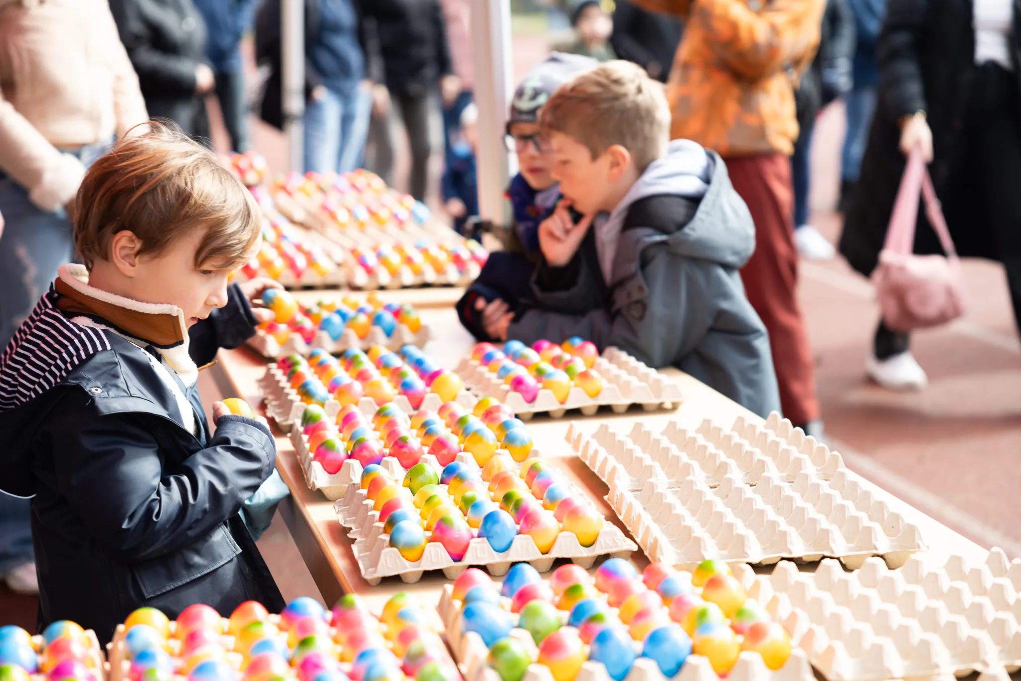 Zwei Jungen suchen auf einem Tisch nach Ostereiern zwischen vielen bunten Eiern in Papphaltern. Im Hintergrund sind weitere Menschen zu sehen.
