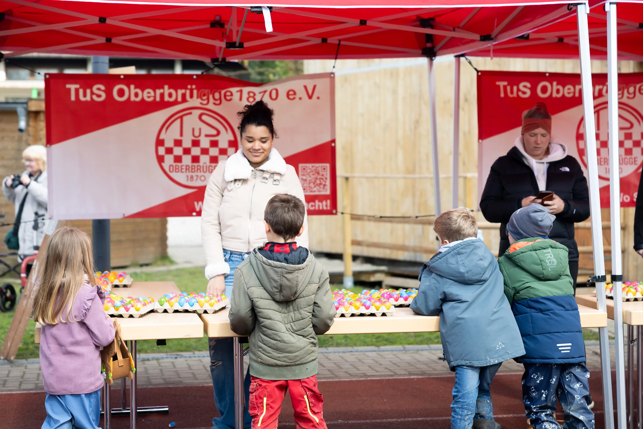 Kinder sammeln unter Aufsicht von Erwachsenen bunte Ostereier auf einem roten Untergrund unter einem roten Pavillon mit dem Logo des TuS Oberbrügge 1870 e.V.