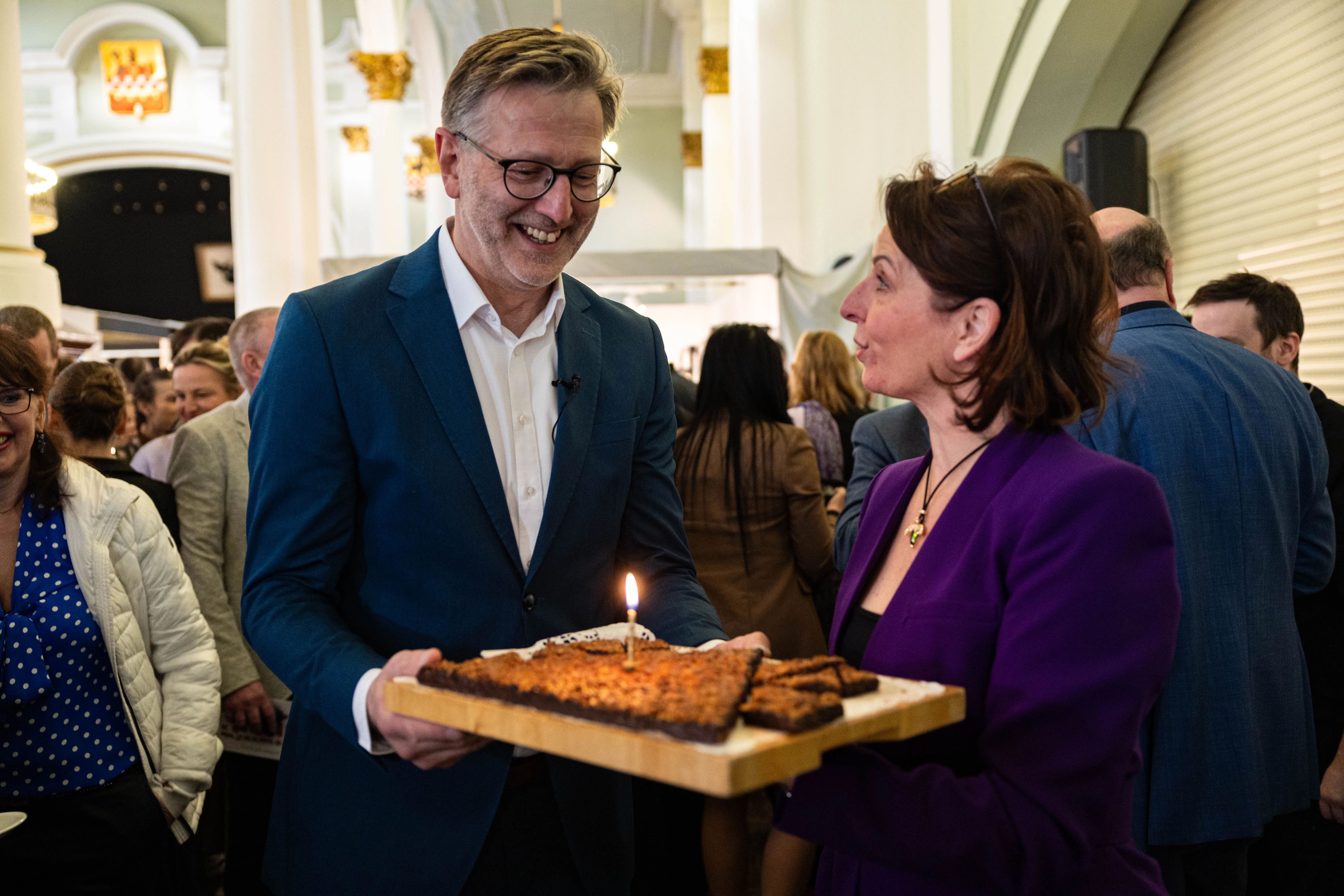 Ein Mann in einem blauen Anzug überreicht einer Frau eine Platte mit Nussecken, auf denen eine einzelne Kerze brennt. Im Hintergrund stehen weitere Menschen in einer hellen Halle.