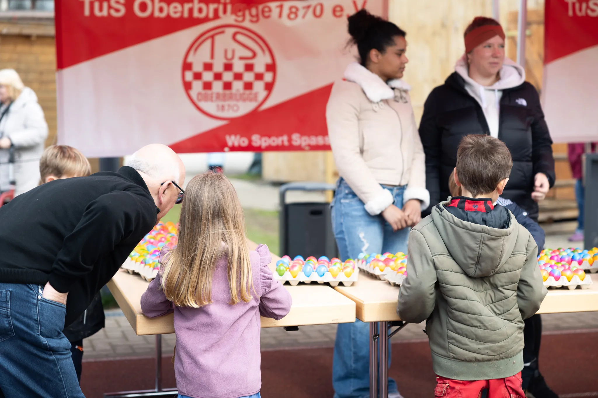 Kinder suchen auf einem Tisch nach Ostereiern, im Hintergrund stehen weitere Personen und ein Banner des TuS Oberbrügge 1870.