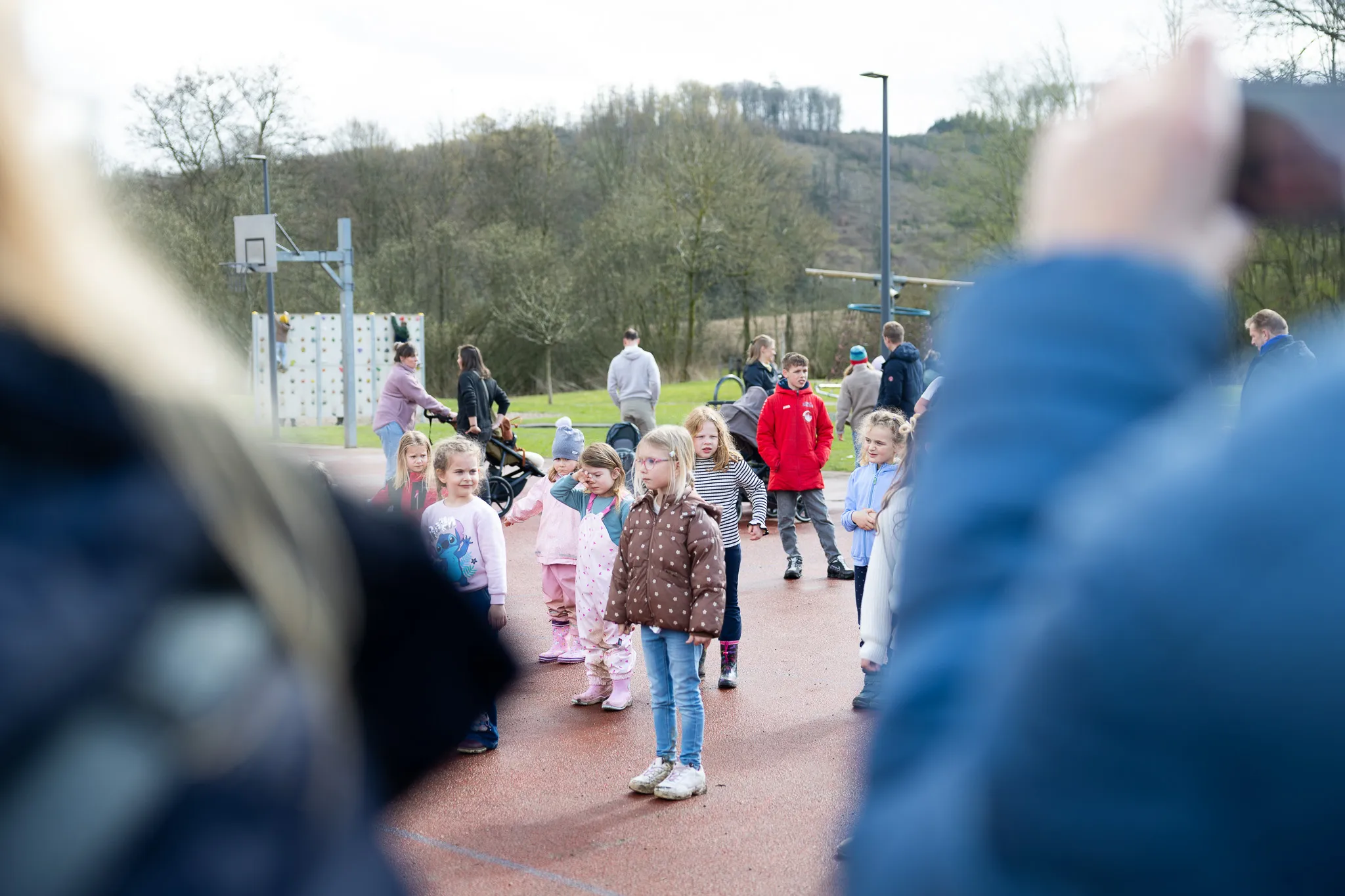 Kinder stehen auf einem roten Belag und suchen nach Ostereiern, während Eltern im Hintergrund zusehen. Im Hintergrund sind Bäume und ein Basketballkorb zu erkennen.