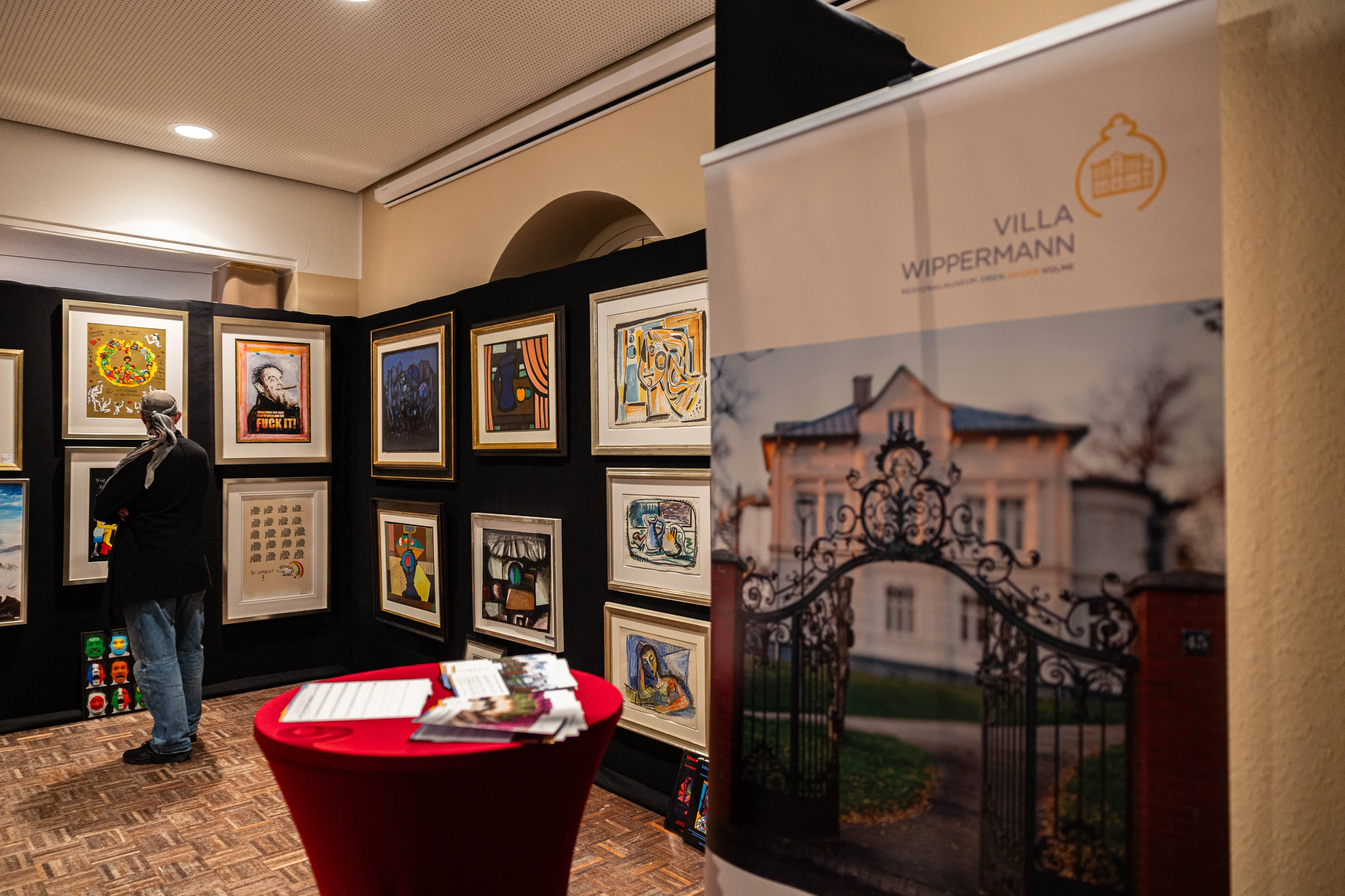 Ausstellungsbesucher betrachtet Gemälde in einem Raum mit dunklen Wänden. Im Hintergrund ein Banner mit dem Bild der Villa Wippermann und dem Hinweis auf eine Ausstellung von Detlev Kümmel.