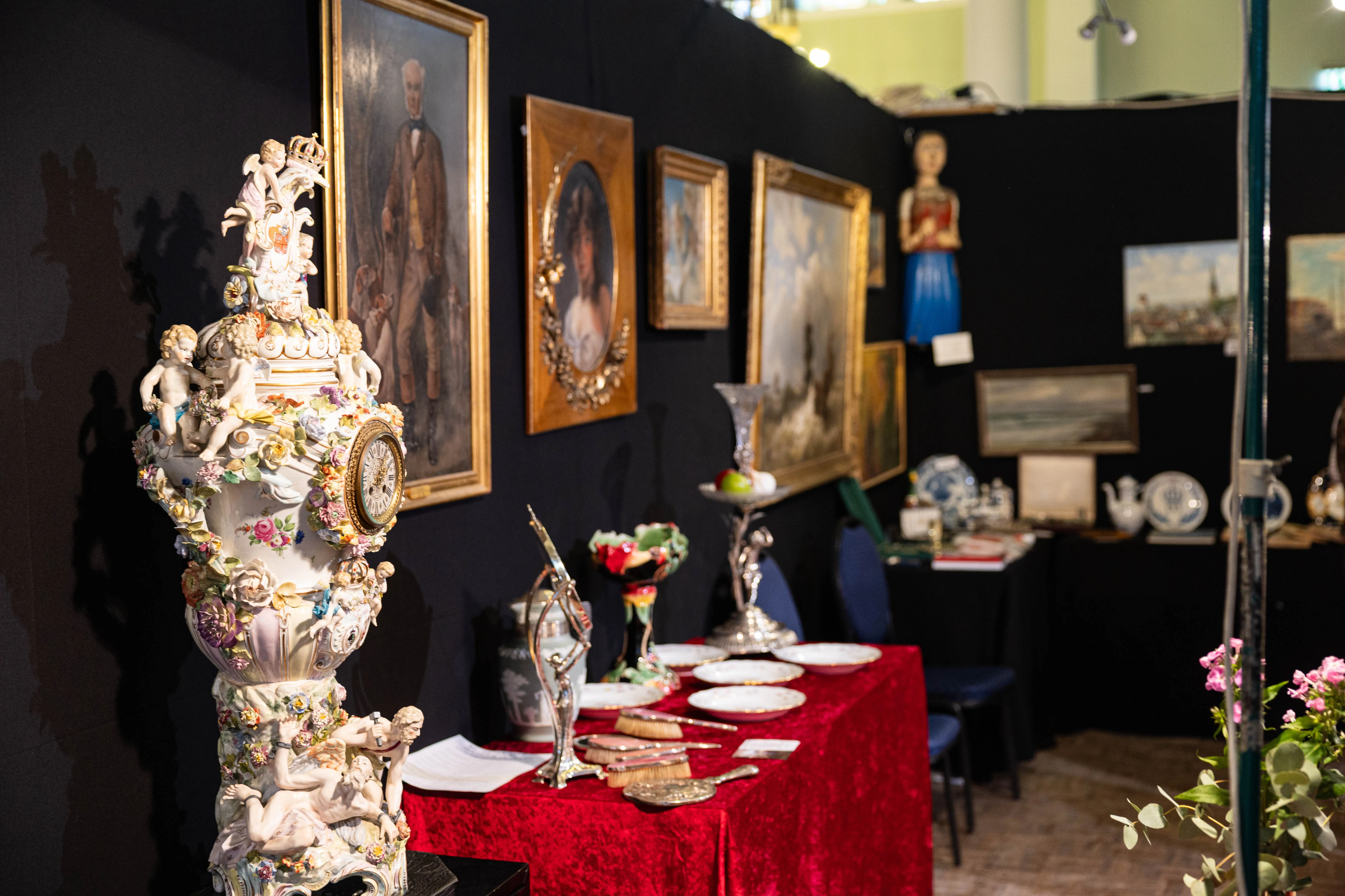 Ausstellung von Kunst und Antiquitäten mit Gemälden, Porzellan und Silberbesteck auf einem roten Tisch.