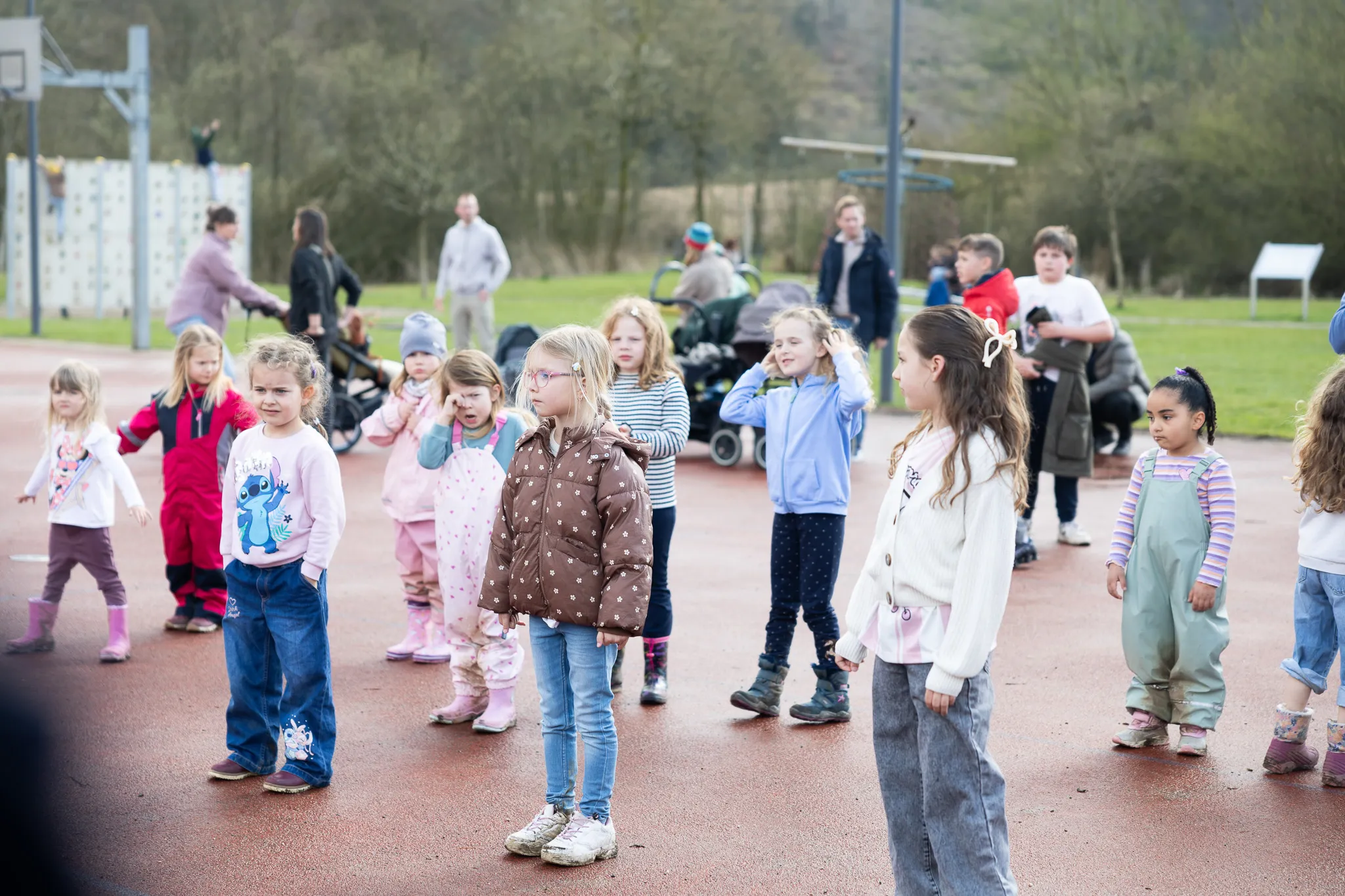 Eine Gruppe Kinder steht auf einem roten Belag und blickt erwartungsvoll in verschiedene Richtungen, während im Hintergrund Erwachsene zusehen. Im Hintergrund sind Bäume und ein Spielplatz zu erkennen.