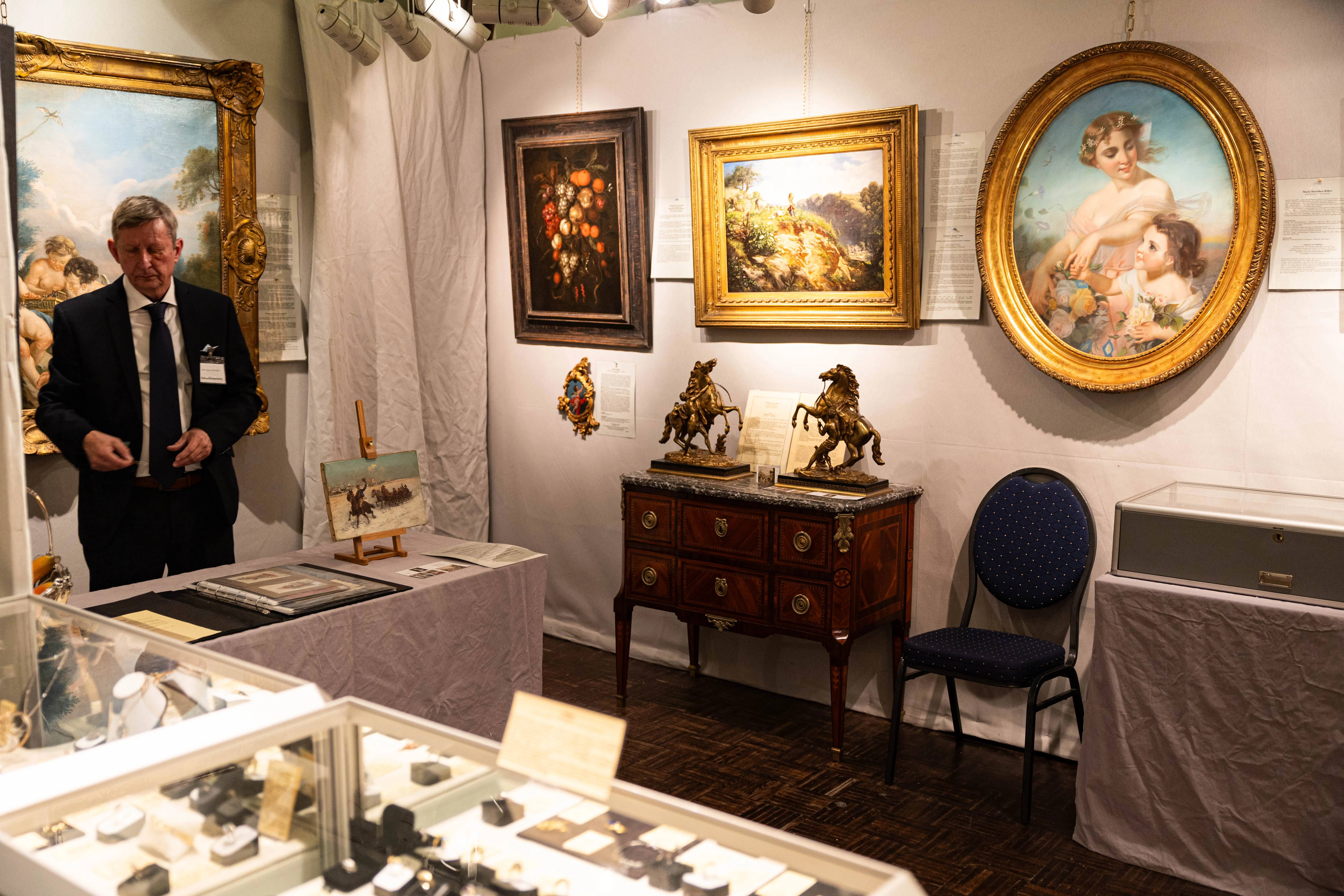 Ein Mann in Anzug und Krawatte steht vor Gemälden und Antiquitäten in einem hellen Ausstellungsraum. Im Vordergrund sind Schmuckstücke in Vitrinen zu sehen.