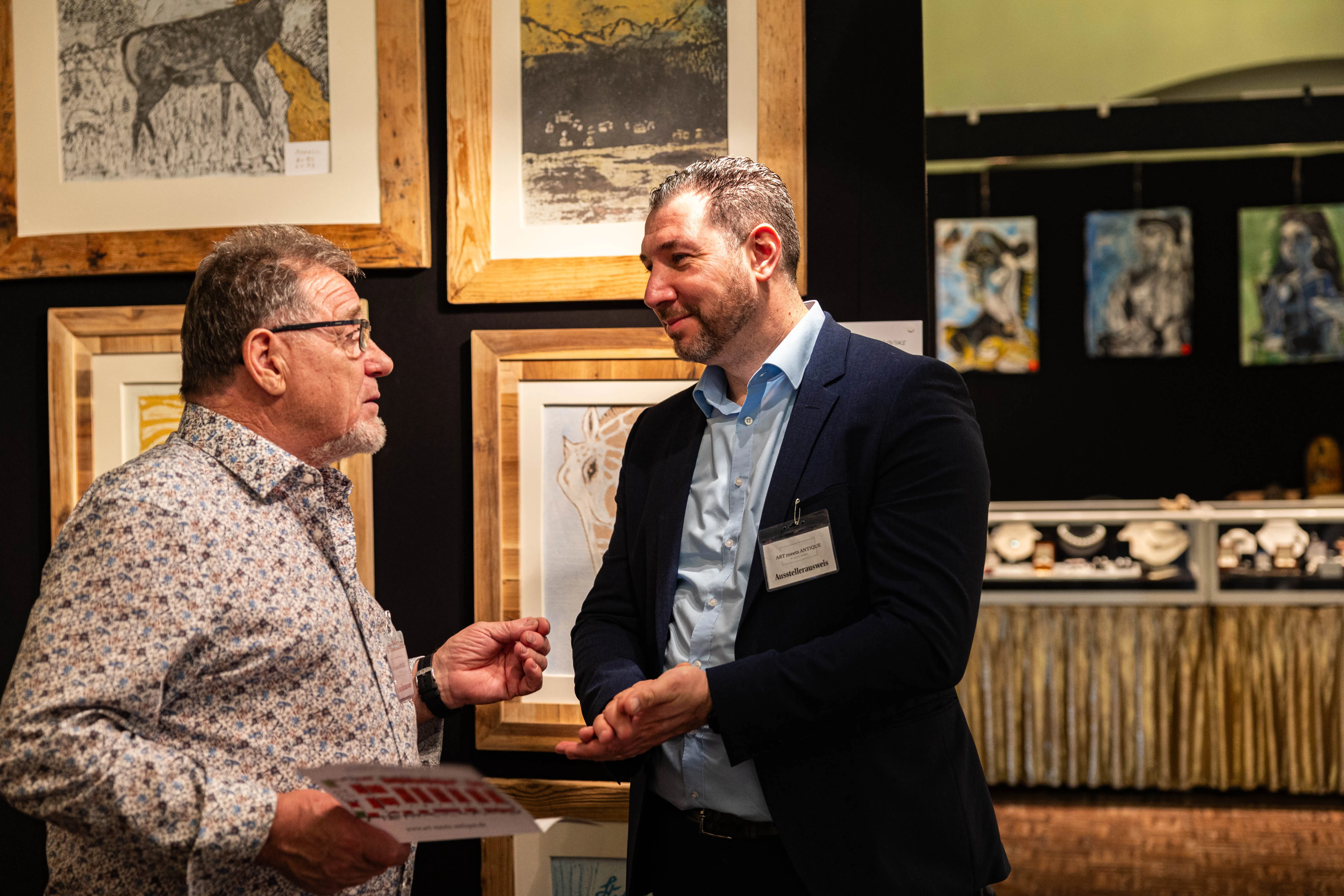 Zwei Männer unterhalten sich auf einer Kunstausstellung. Im Hintergrund hängen gerahmte Bilder an den Wänden.