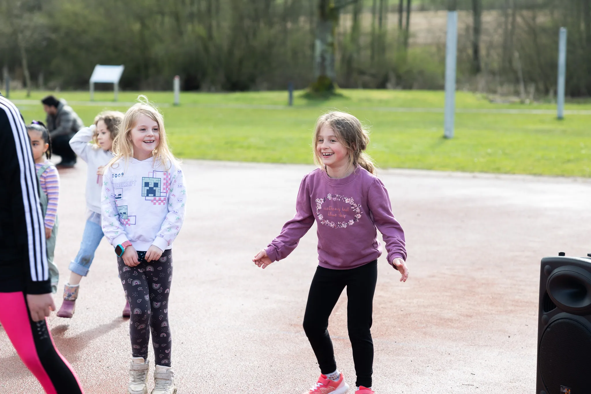 Zwei Mädchen freuen sich während einer Ostereiersuche im Freien. Im Hintergrund sind weitere Kinder und Bäume zu sehen.