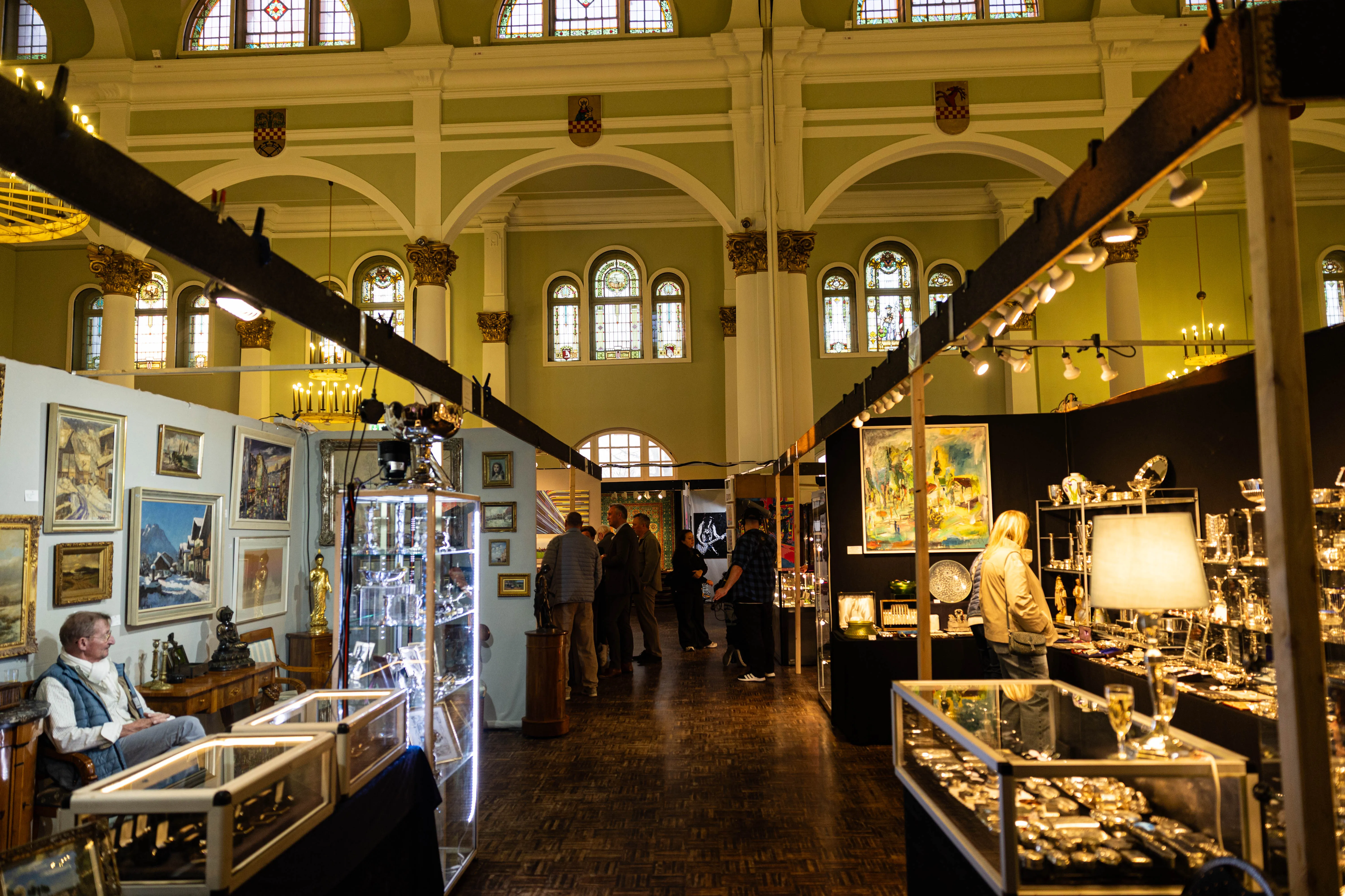 Innenansicht einer Kunstausstellung mit Besuchern vor Gemälden und Antiquitätenständen in einem hellen Raum mit hohen Decken und Bogenfenstern.