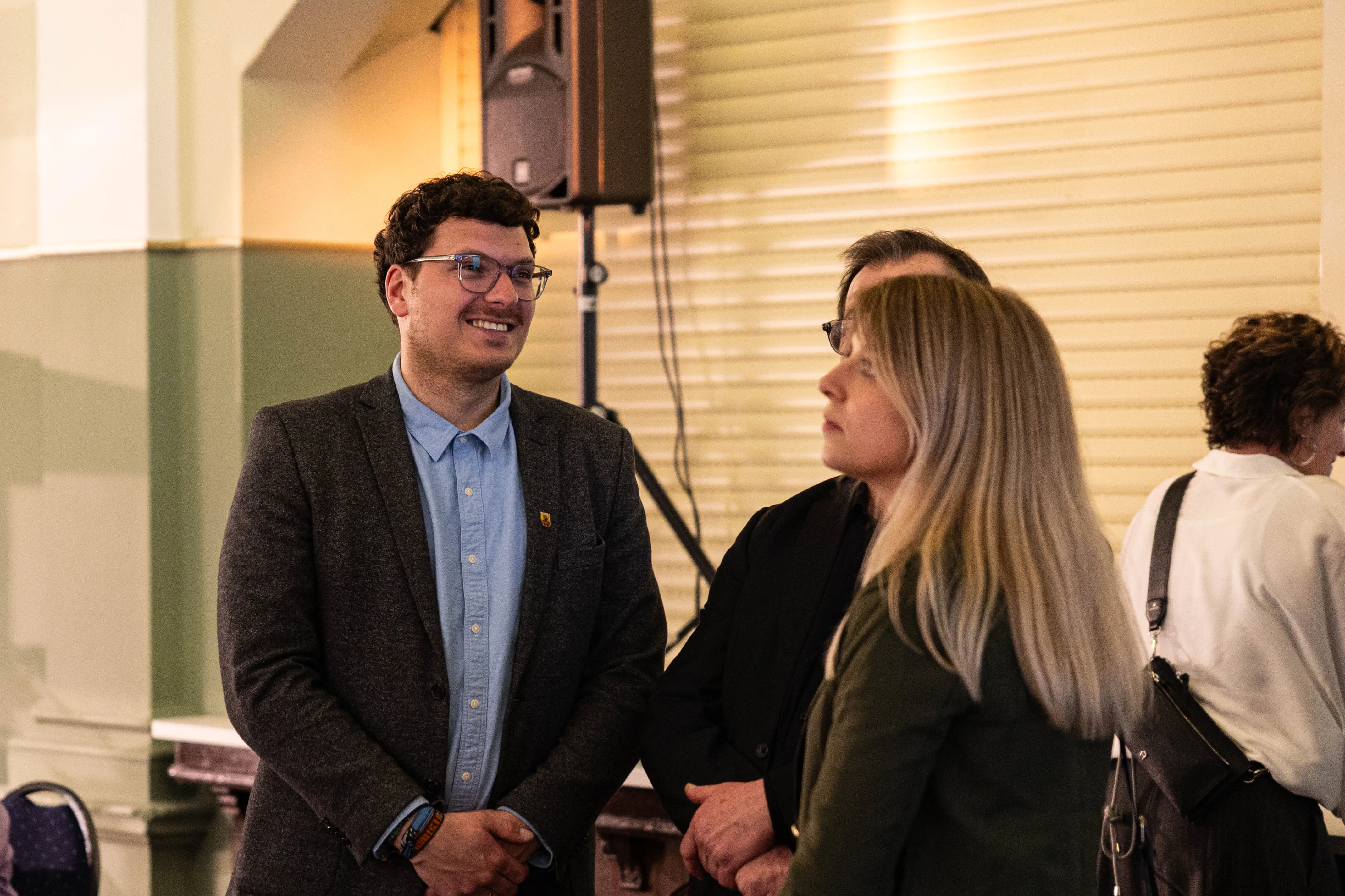 Dominik Hass-Sommer, ein Mann mit dunklem lockigem Haar und Brille, unterhält sich auf einer Vernissage mit einer Frau.