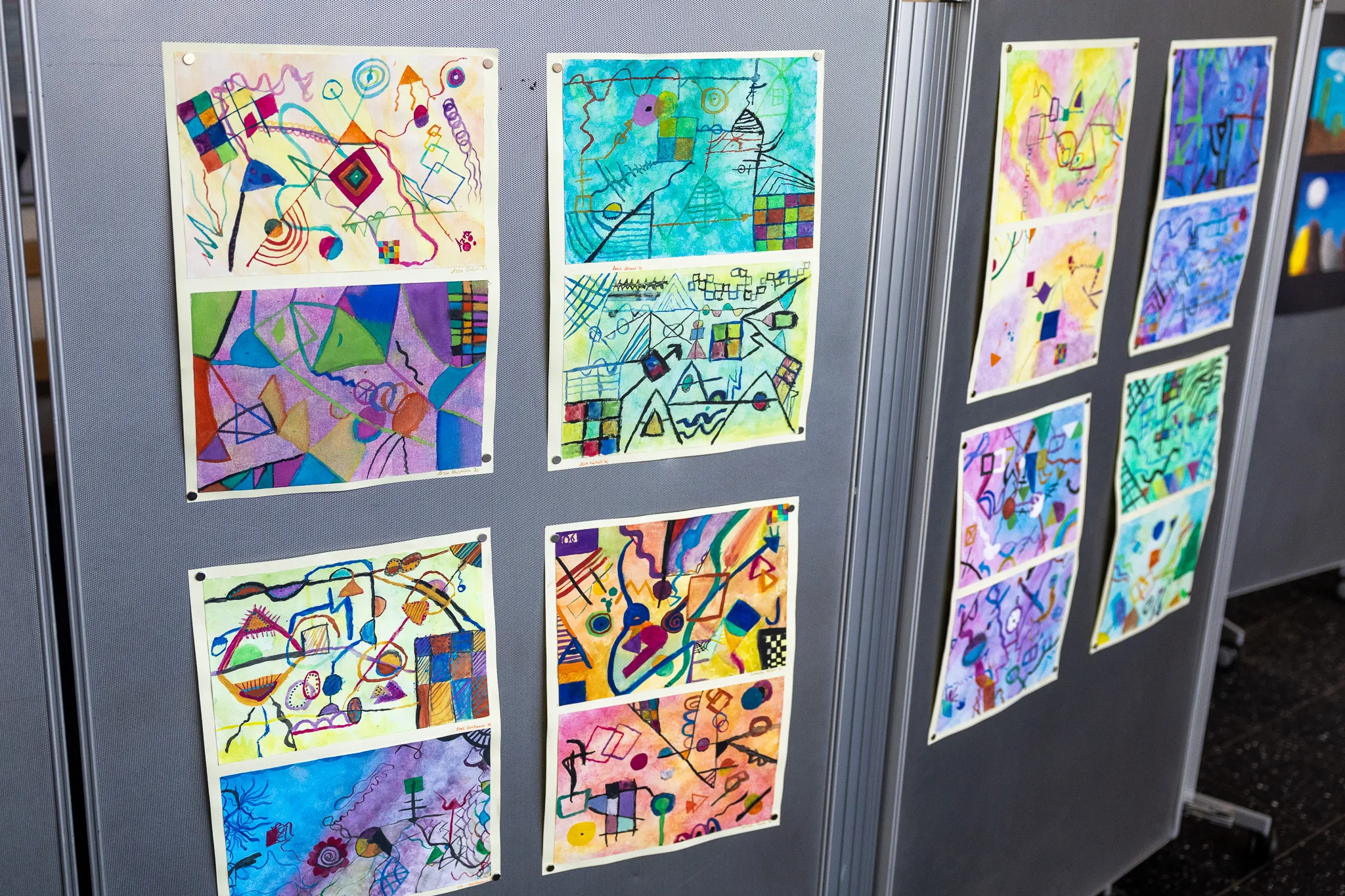 Sechs farbenfrohe Kinderzeichnungen mit geometrischen Formen und abstrakten Mustern sind an einer grauen Stellwand befestigt.